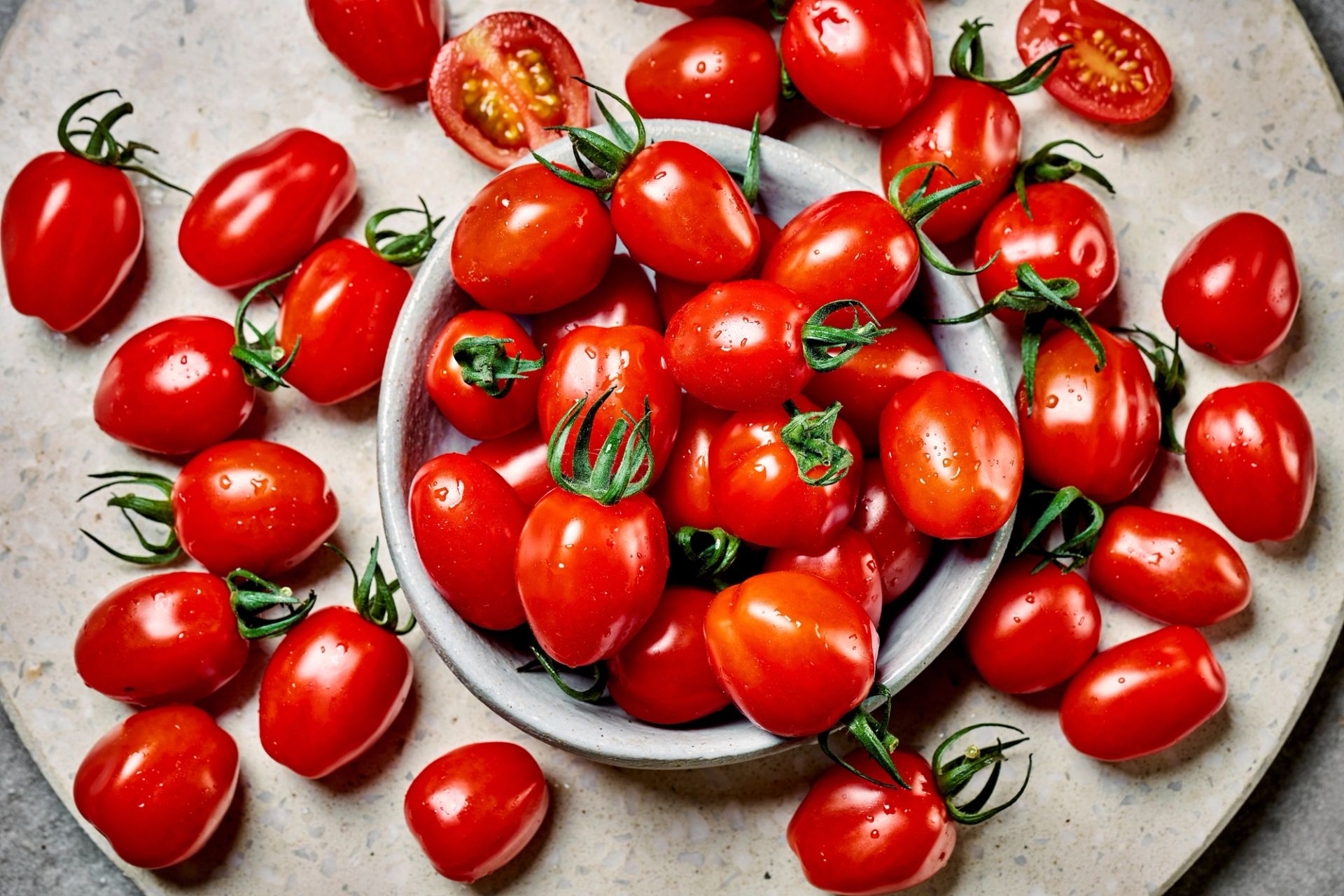 Skål fylt med røde plommetomater står på et lyst underlag, omgitt av flere tomater spredt rundt. Noen av tomatene er delt i to og viser det saftige fruktkjøttet, mens vanndråper på skallet fremhever friskheten.