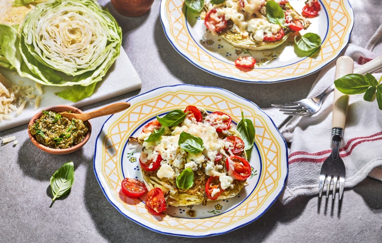 To tallerkener med ovnsbakt kål toppet med cherrytomater, smuldret ost og friske basilikumblader, servert ved siden av et halvt kålhode og en skål med grønn pesto på et grått bord.