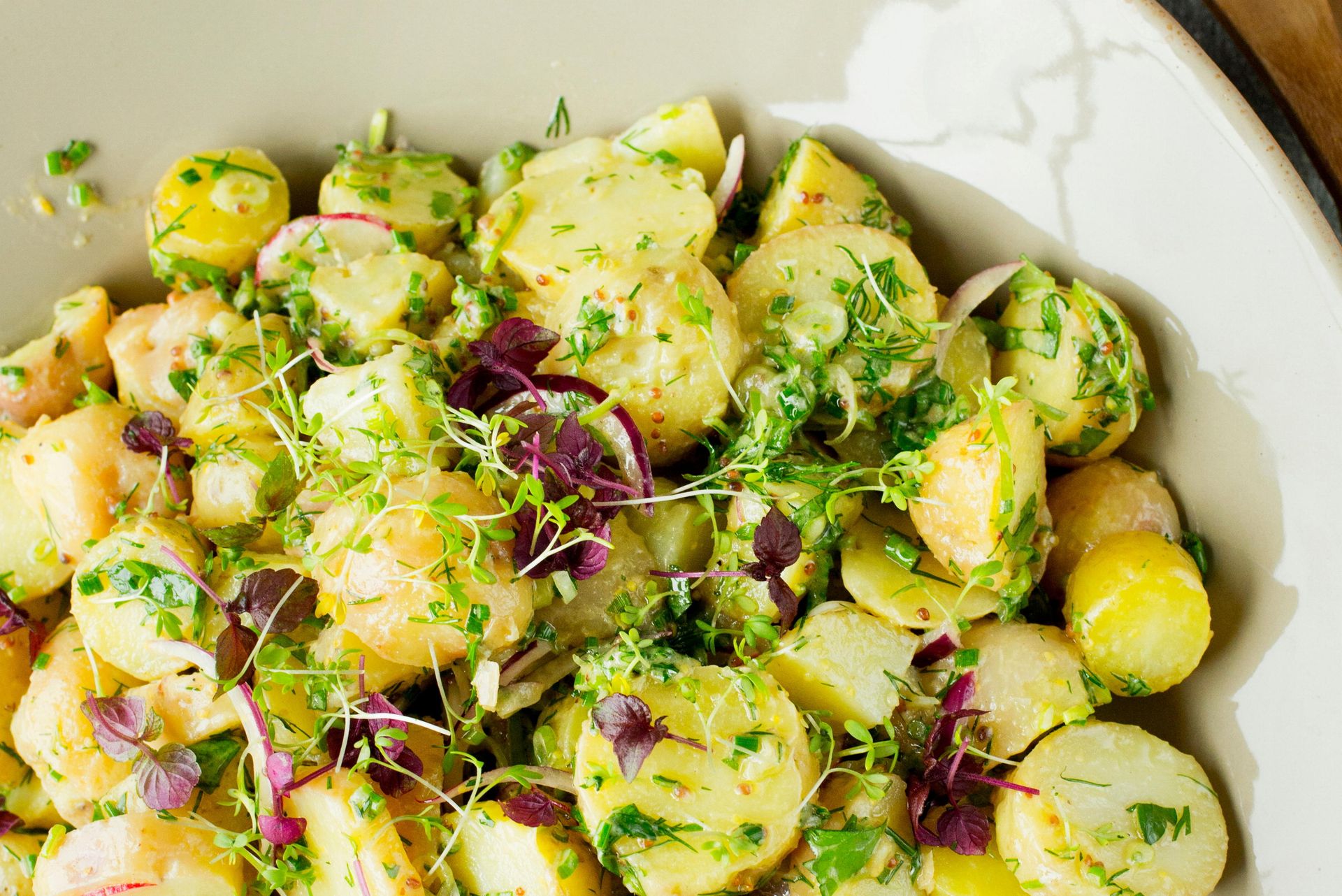 En lys, rund tallerken fylt med potetsalat laget av små poteter i skiver. Potetene er blandet med friske urter som persille og dill, samt tynne skiver av rødløk. Salaten er lett dekket av en grønn, urtepregt dressing, og noen spirer ligger drysset på toppen. I bakgrunnen ligger to tresleiver delvis synlige.