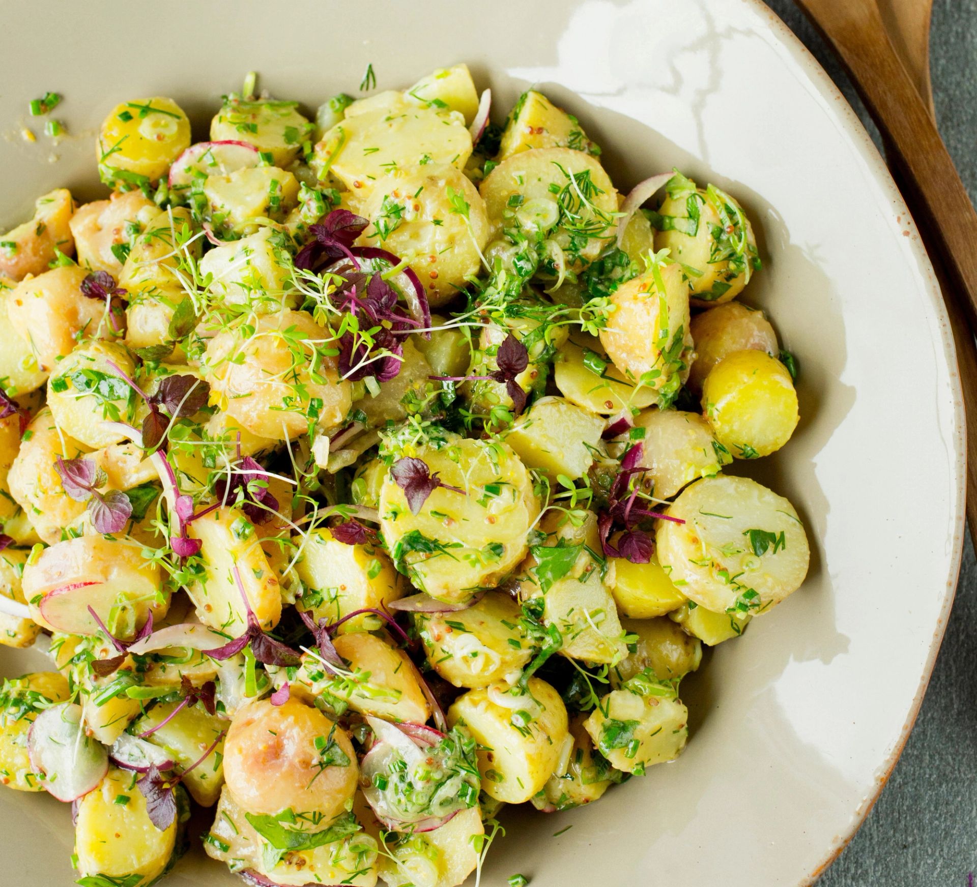 En lys, rund tallerken fylt med potetsalat laget av små poteter i skiver. Potetene er blandet med friske urter som persille og dill, samt tynne skiver av rødløk. Salaten er lett dekket av en grønn, urtepregt dressing, og noen spirer ligger drysset på toppen. I bakgrunnen ligger to tresleiver delvis synlige.
