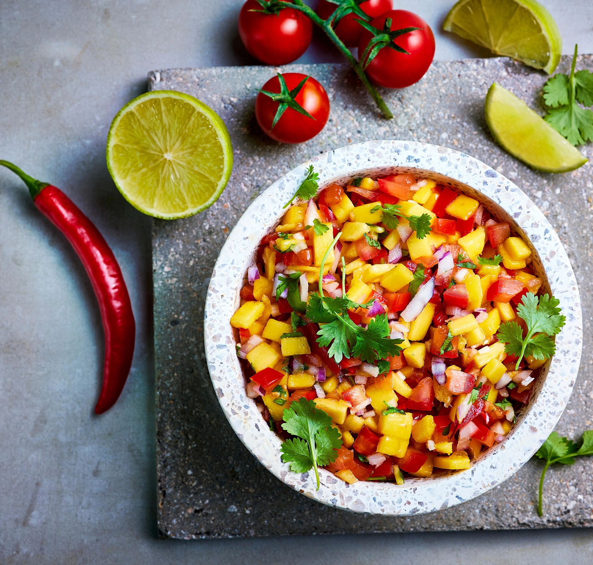 Bolleskål med fruktig mango- og tomatsalsa toppet med koriander, omkranset av limebåter, cherrytomater og en rød chili på grå bakgrunn.