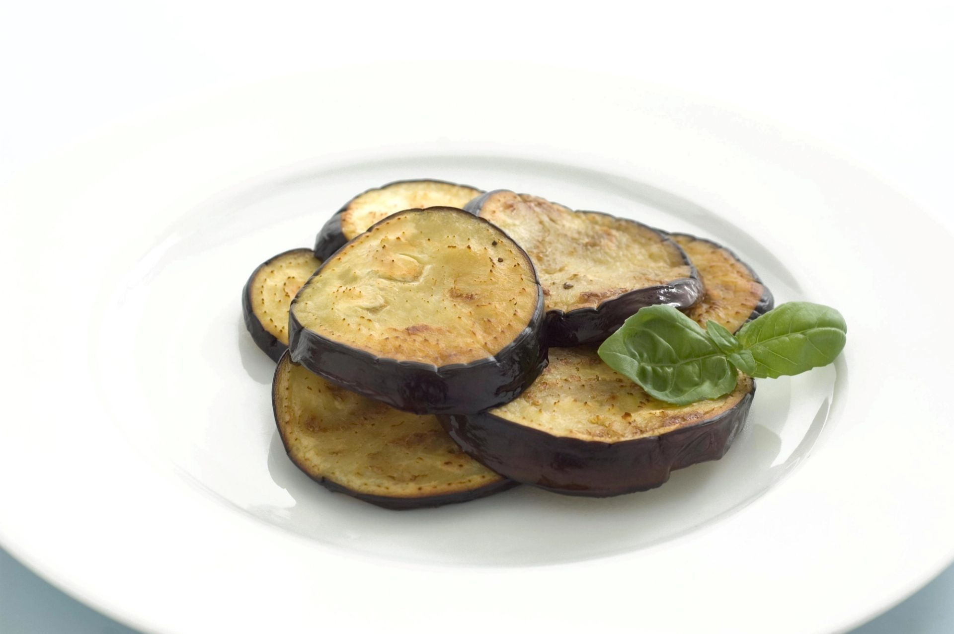 Stekte skiver av aubergine anrettet på hvit tallerken, pyntet med et grønt basilikumblad.