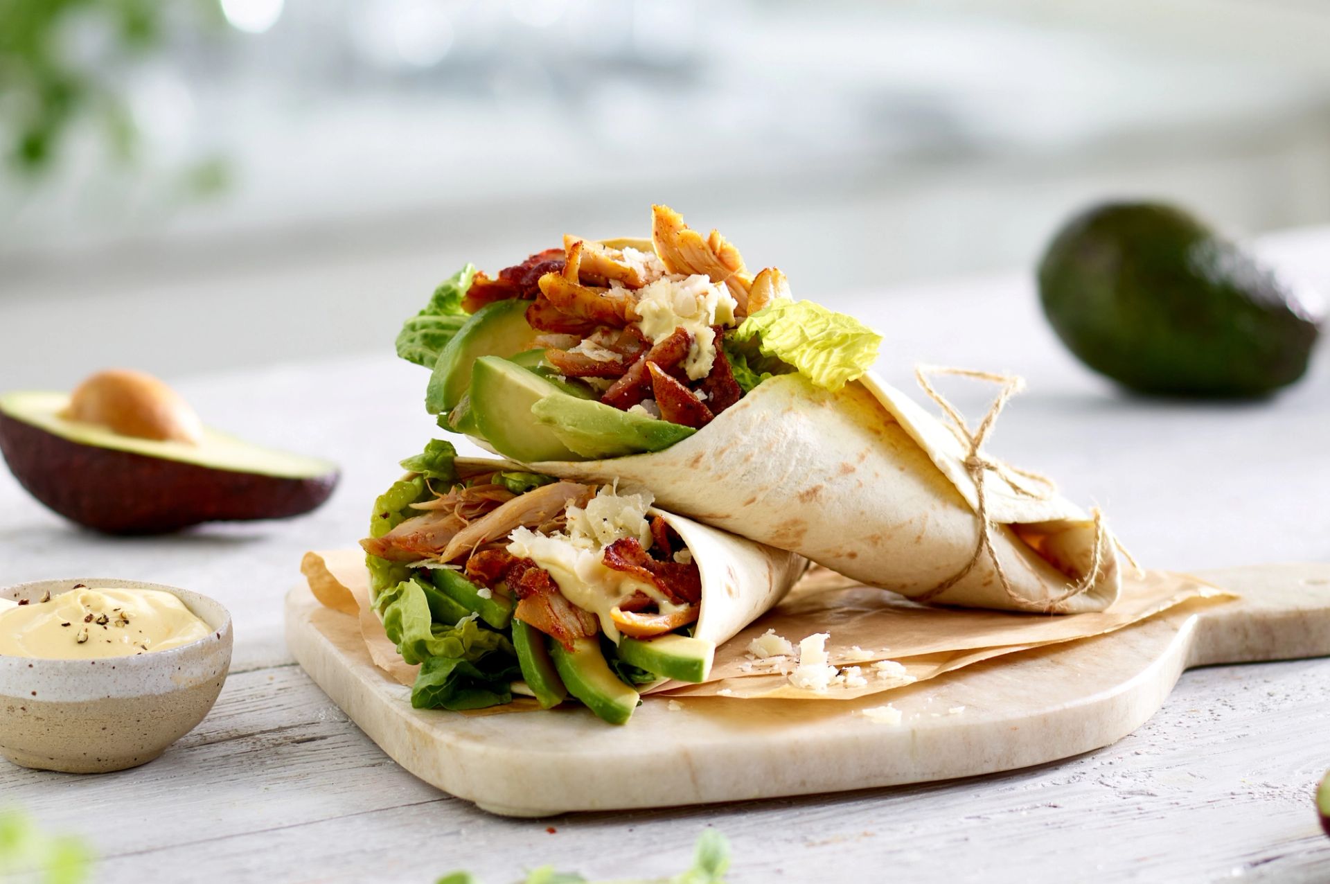 Cæsarsalat-inspirert wrap fylt med avokado, sprø hjertesalat, grillet kylling, bacon og revet parmesan, servert på en lys trefjøl med avokado og dressing i bakgrunnen.