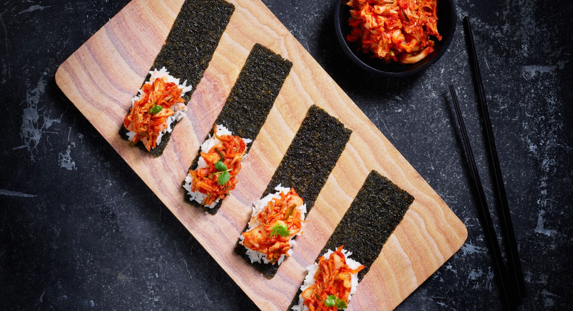 Fire avlange stykker med tang (gim) ligger på et trefat, hver toppet med en porsjon hvit ris og spicy kimchi. Til høyre står en liten skål med ekstra kimchi, og et par svarte spisepinner ligger ved siden av på den mørke overflaten.
