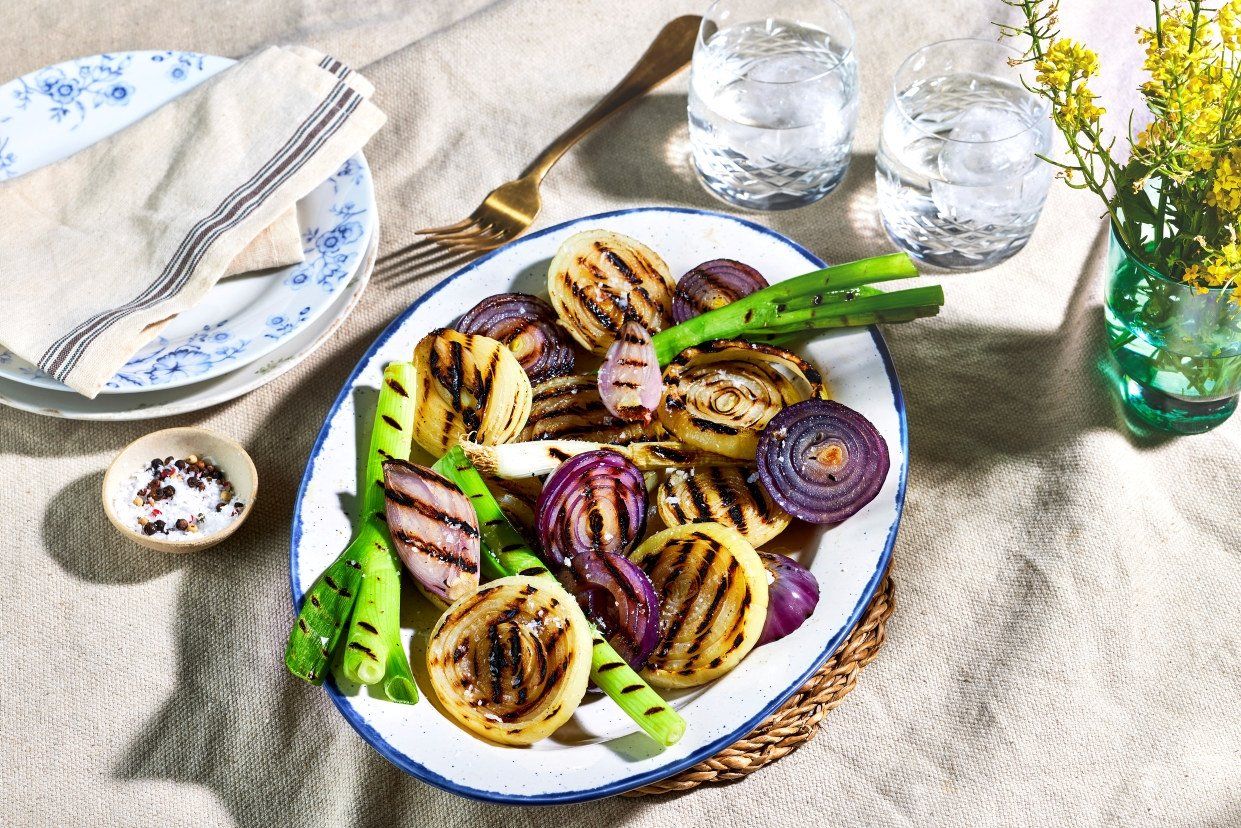 En tallerken med grillede løkringer i gule og lilla nyanser og grønne aspargesbiter, servert på et lyst bord med vannglass, servise og en liten blomsterbukett ved siden av.