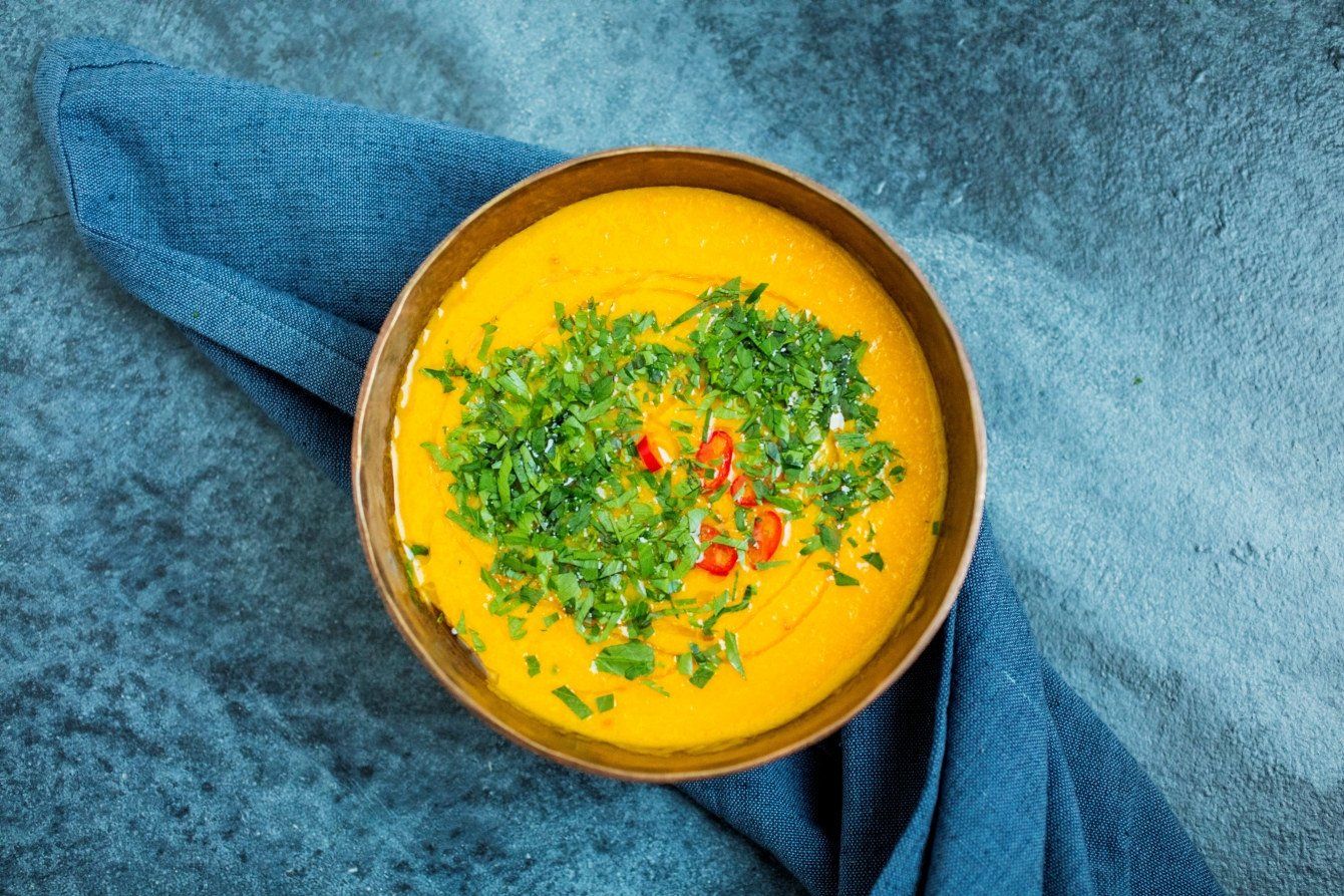 Skål med kremet gulrotsuppe toppet med frisk koriander og skiver av rød chili. Skålen står på en blå duk med blå serviett.