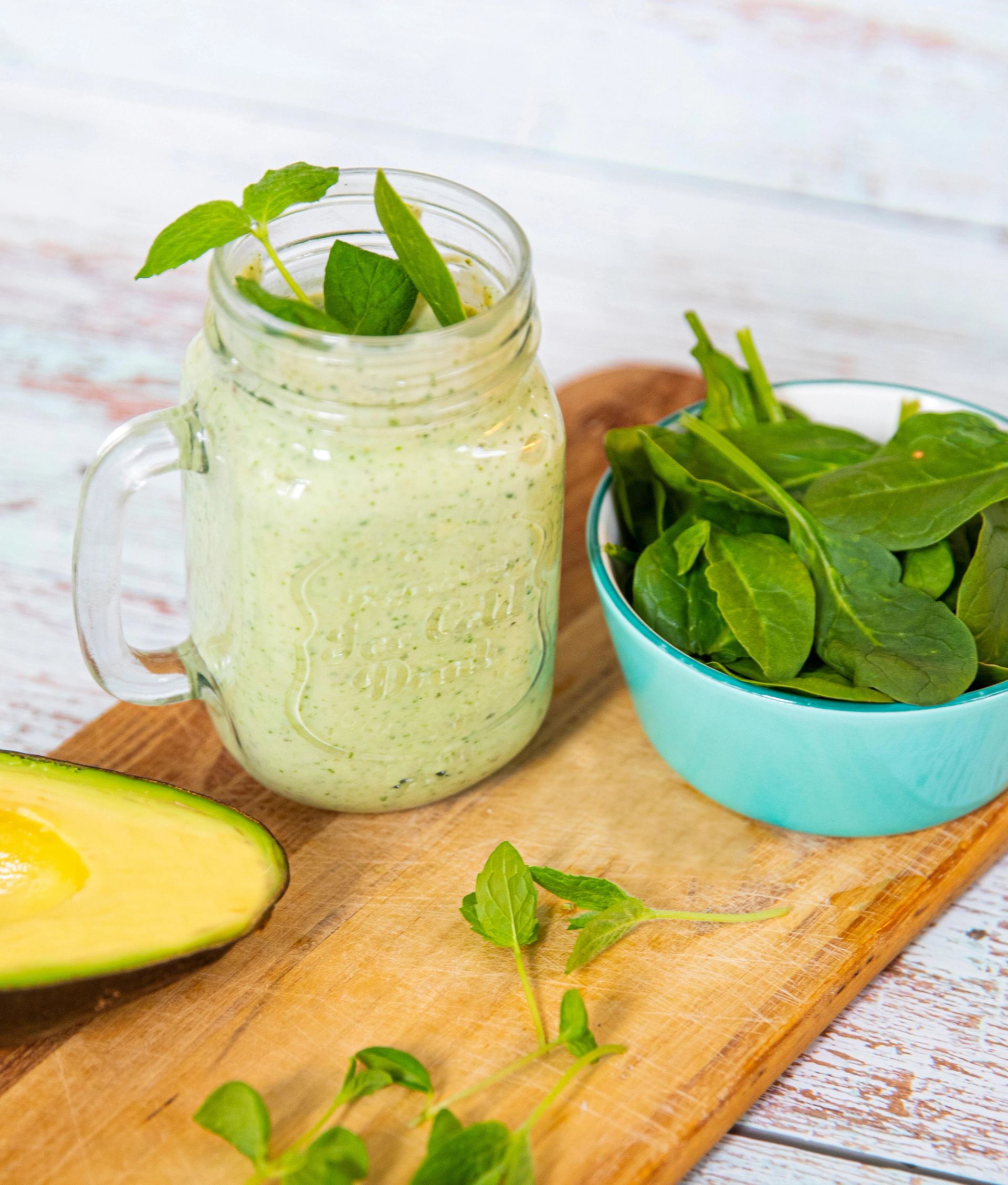 Grønn smoothie i glasskrus med spinat og mynte, ved siden av en bolle med spinat og en halv avokado.