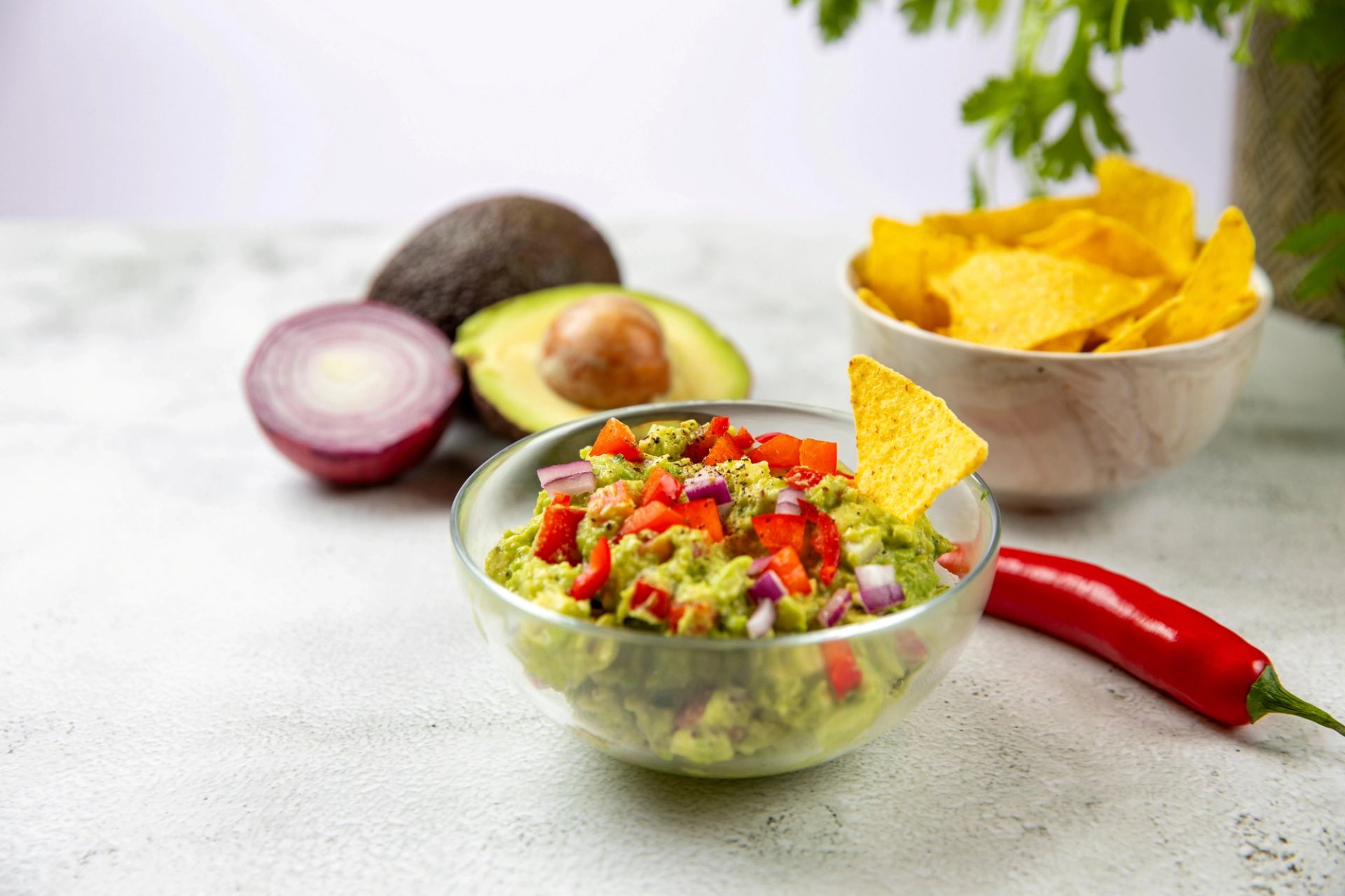 Skål med guacamole toppet med rød paprika og rødløk, servert med tortillachips. Avokado, rødløk og chili ligger i bakgrunnen.