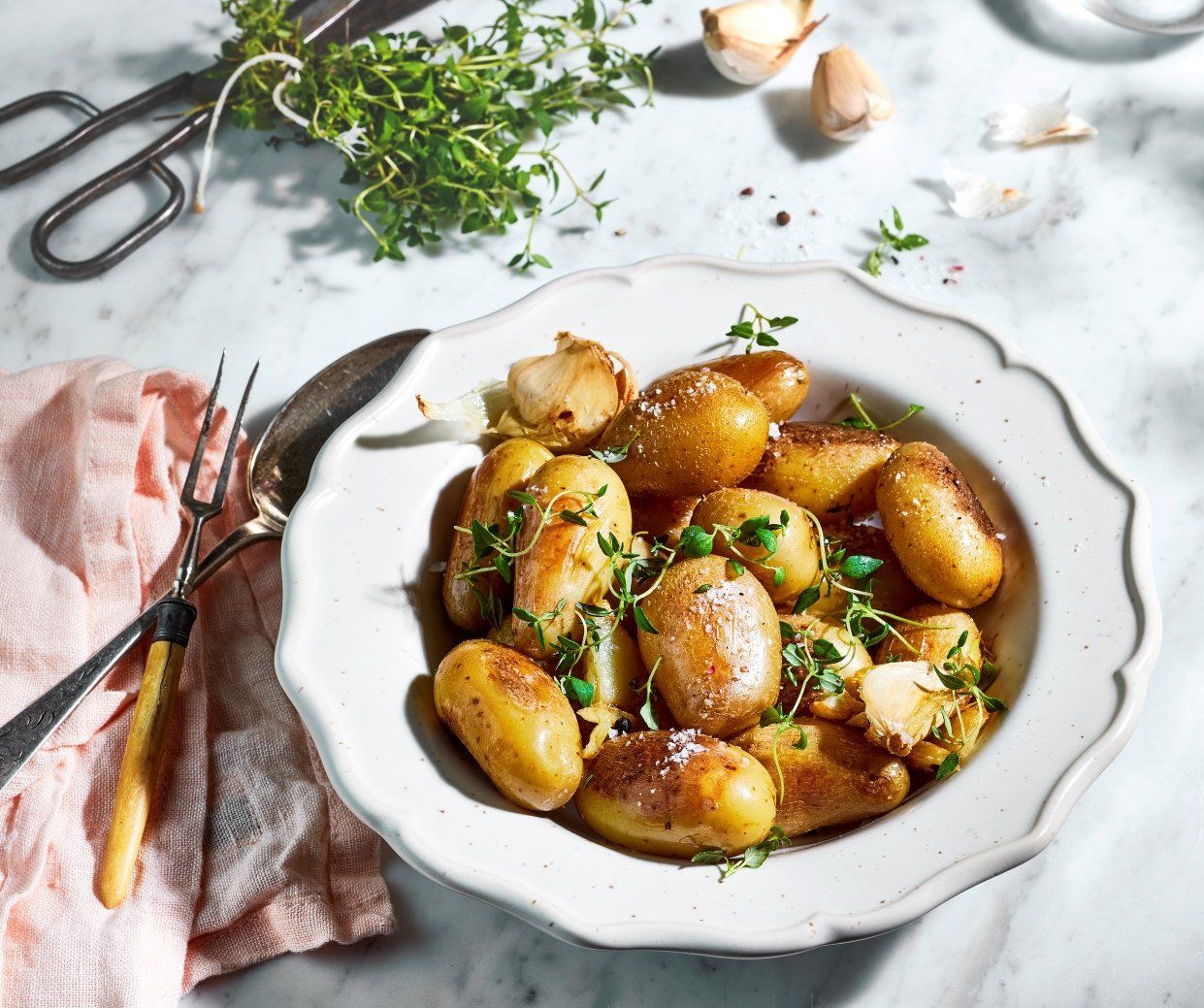 En hvit, mønstret dyp tallerken fylt med gylne, ovnsbakte småpoteter. Potetene er toppet med fersk timian, flaksalt og bakte hvitløksfedd.
