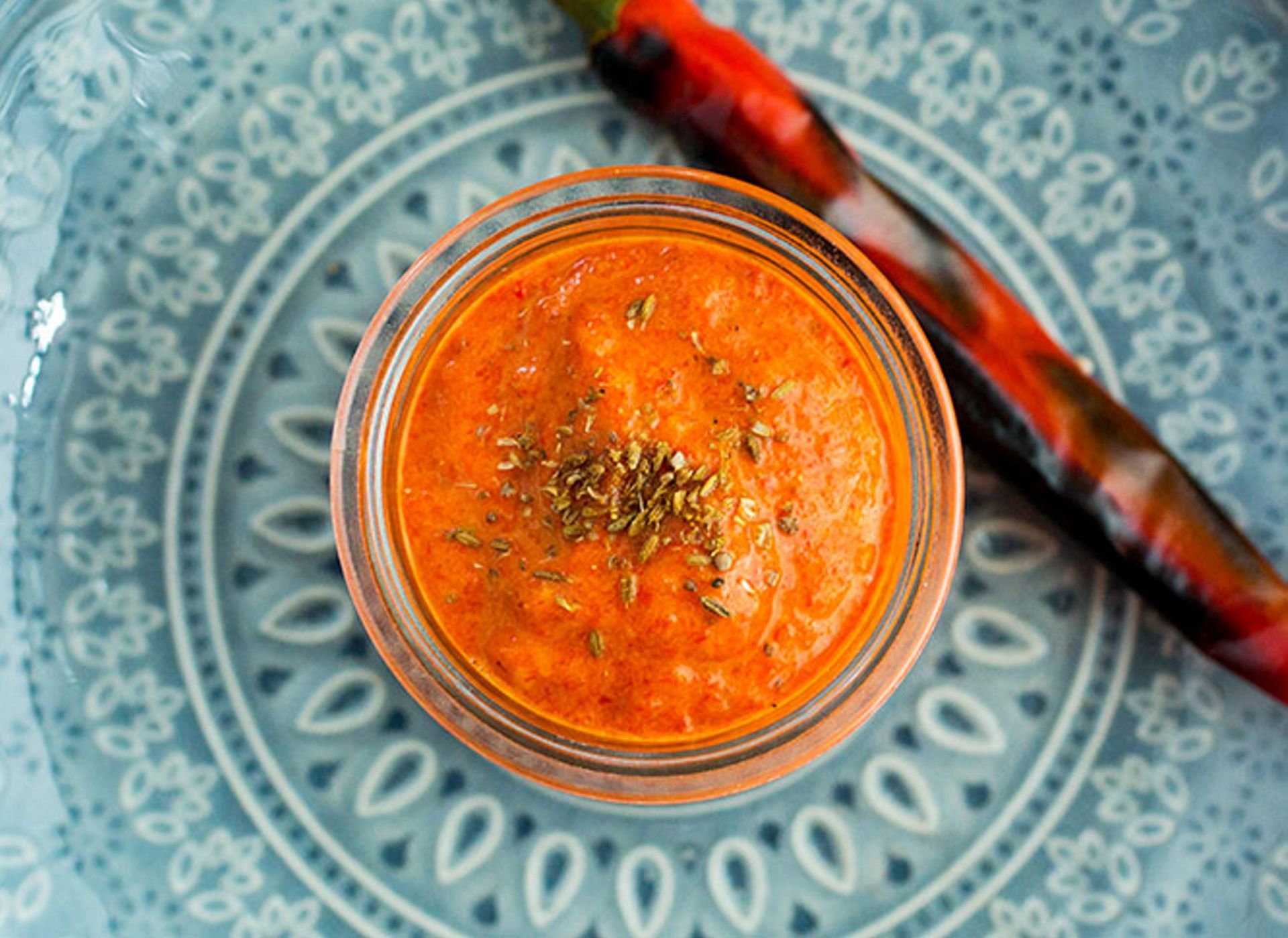 Harissa i et glass, drysset med tørkede urter, servert på en blå mønstret tallerken med røde chilipepper ved siden av.