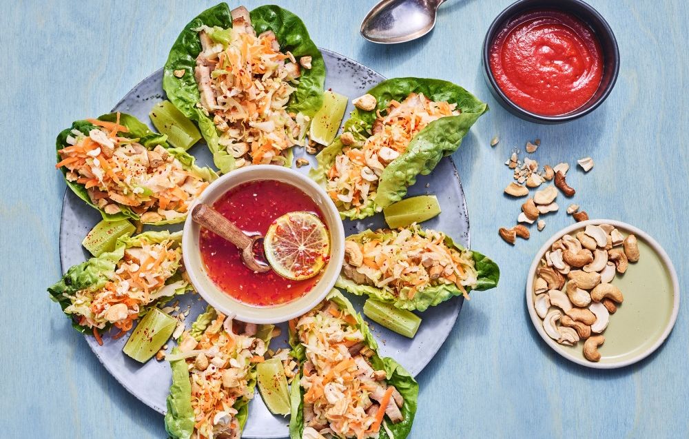 Hjertesalatblader fylt med strimlet svineribbe, gulrot og kål, cashewnøtter og en rød dippsaus på et stort fat.