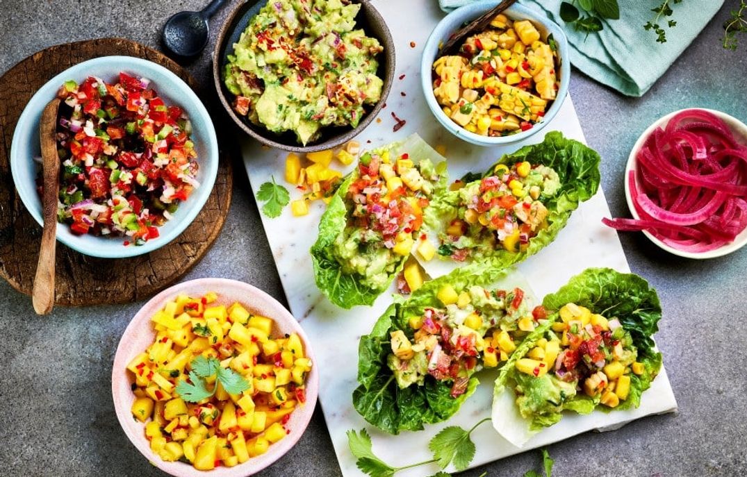 Salatwraps av hjertesalat fylt med guacamole, mango- og tomatsalsa, servert på et fat omgitt av skåler med salsa, syltet rødløk og ekstra mangosalat på et grått bord.