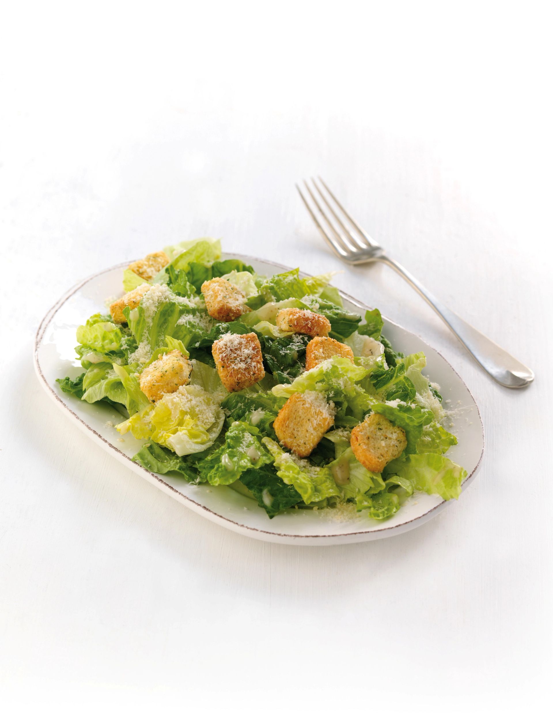 Caesarsalat med romanosalat, krutonger og revet parmesan servert på hvit tallerken, med gaffel ved siden av.