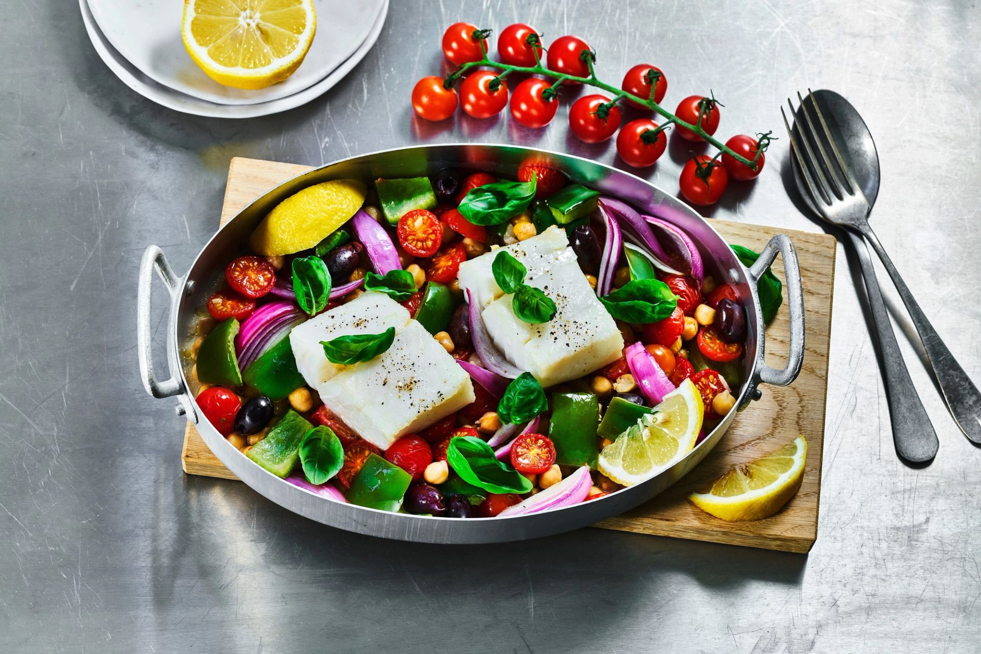 En ovnsform med bakt hvit fisk servert med cherrytomater, paprika, rødløk, kikkerter, oliven og fersk basilikum, toppet med sitronskiver på et bord.