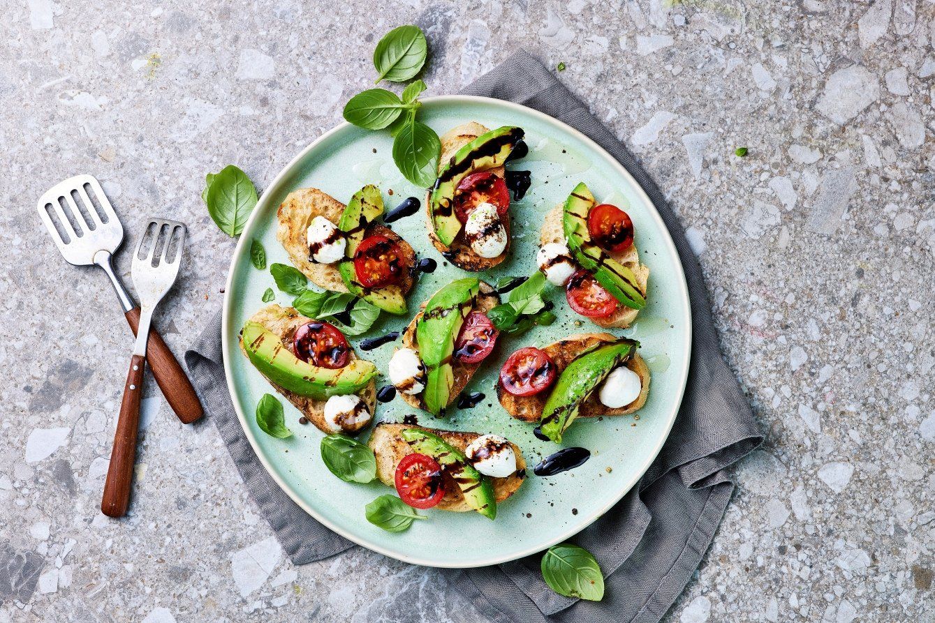 Crostini toppet med grillet avokado, cherrytomater, mozzarella, basilikum og balsamico, servert på en lys grønn tallerken liggende på en grå steinoverflate.