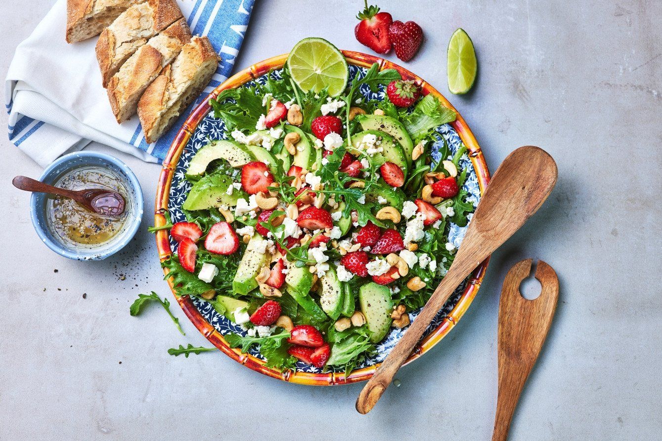 Fargerik sommersalat med avokado, jordbær, ruccula, fetaost og cashewnøtter i en dekorativ bolle, servert med lime og brød ved siden av.