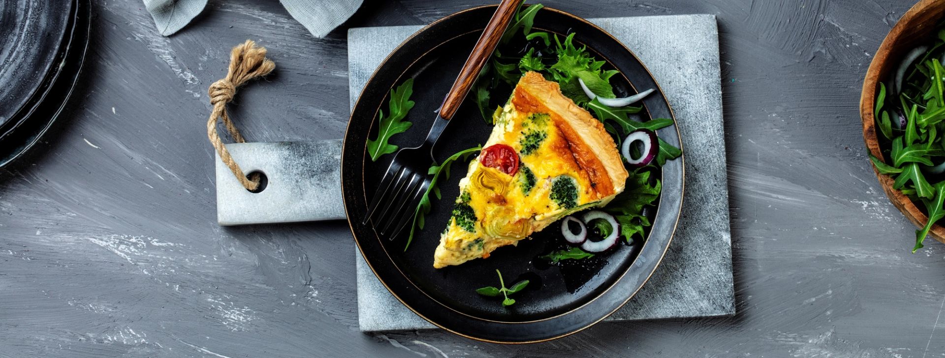 En sort tallerken med et kakestykke av quiche med gyllen skorpe, synlige biter av broccoli, tomat og løk. Quichen ligger på en seng av grønne bladgrønnsaker sammen med ringer av rødløk. Ved siden av ligger en svart gaffel med treskaft. Tallerkenen står på en lys steinplate mot en mørkgrå bakgrunn.