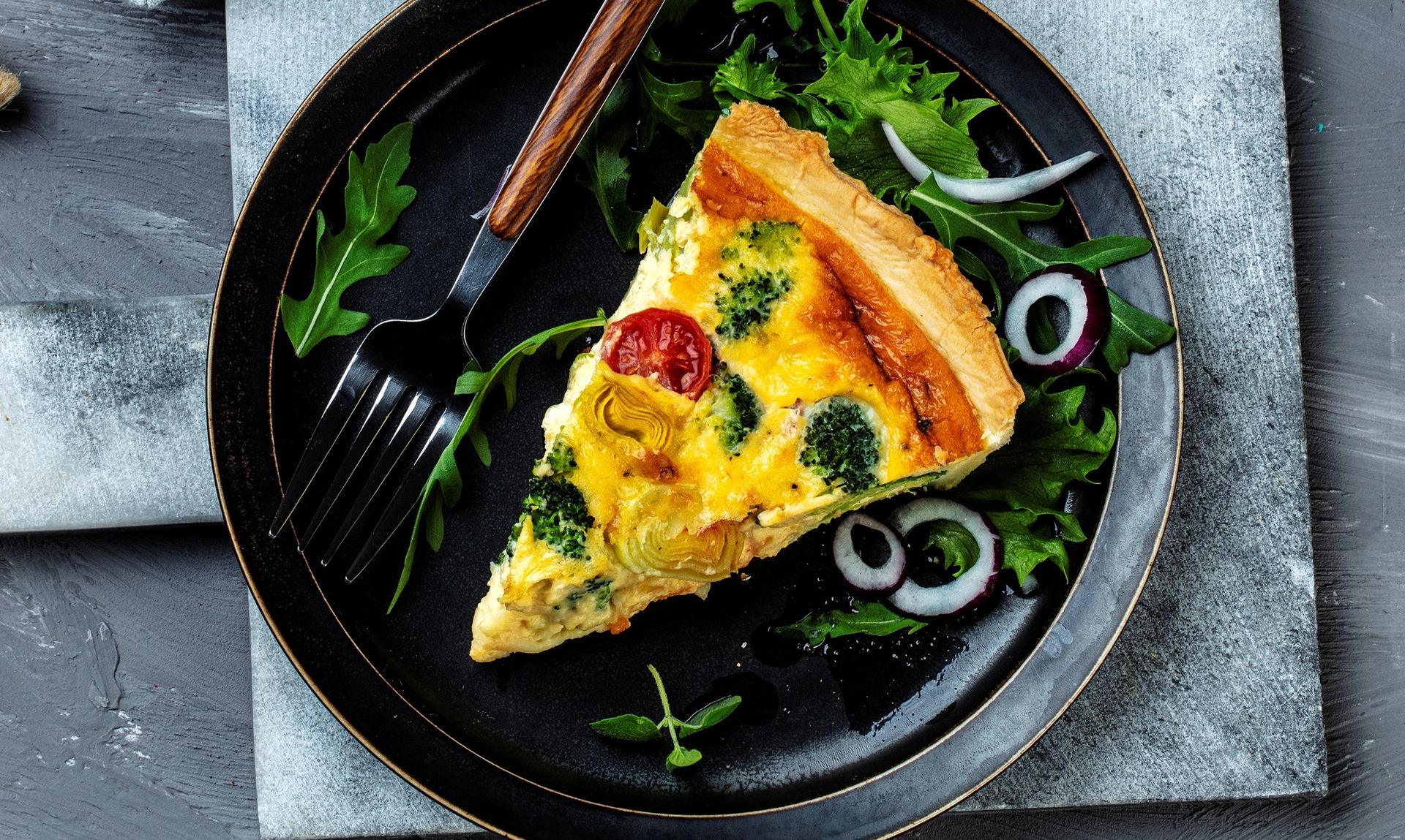 En sort tallerken med et kakestykke av quiche med gyllen skorpe, synlige biter av brokkoli, tomat og løk. Quichen ligger på en seng av grønne bladgrønnsaker sammen med ringer av rødløk. Ved siden av ligger en svart gaffel med treskaft. Tallerkenen står på en lys steinplate mot en mørkgrå bakgrunn.