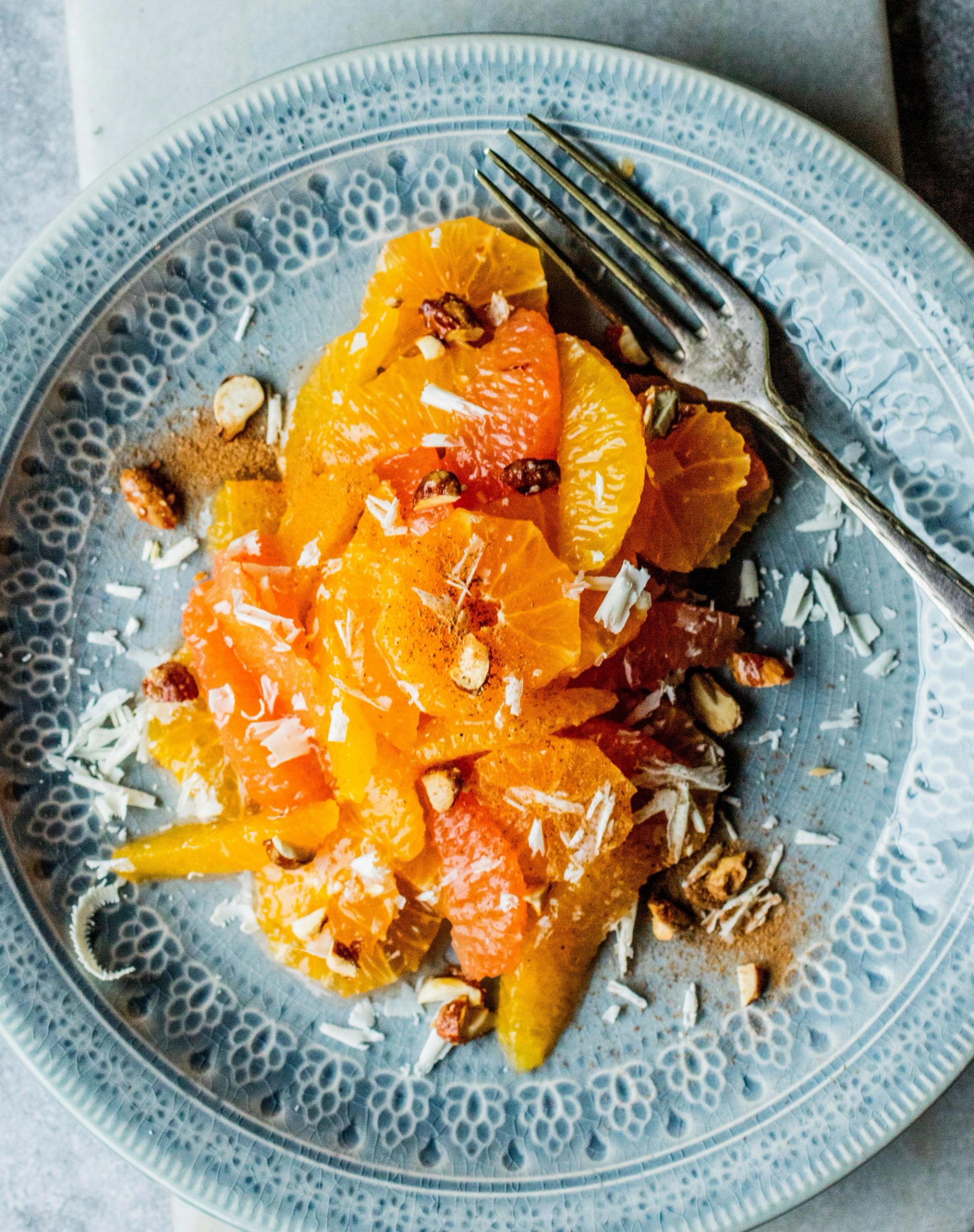 Et fat sett ovenfra med skiver av appelsin og grapefrukt lagt i lag, drysset med hakkede nøtter, kokosflak og krydder, servert på en dekorert blå tallerken med en gaffel plassert øverst.