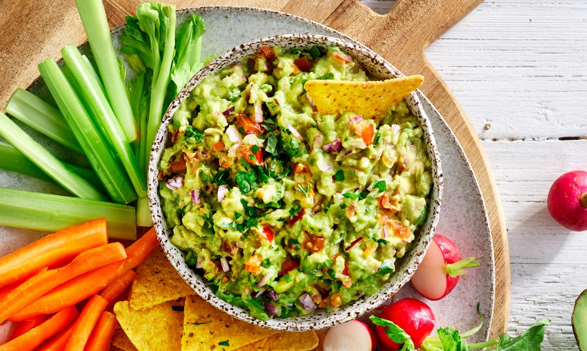 Skål med hjemmelaget guacamole fylt med hakket avokado, tomat, rødløk og koriander, servert med tortillachips, selleristenger og gulrotstaver.