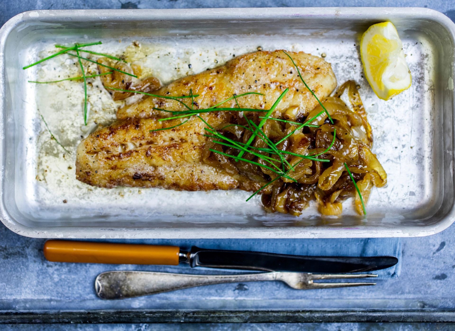 Grillet fiskefilet servert på fat med stekt løk, gressløk og en sitronbåt, med bestikk ved siden av.
