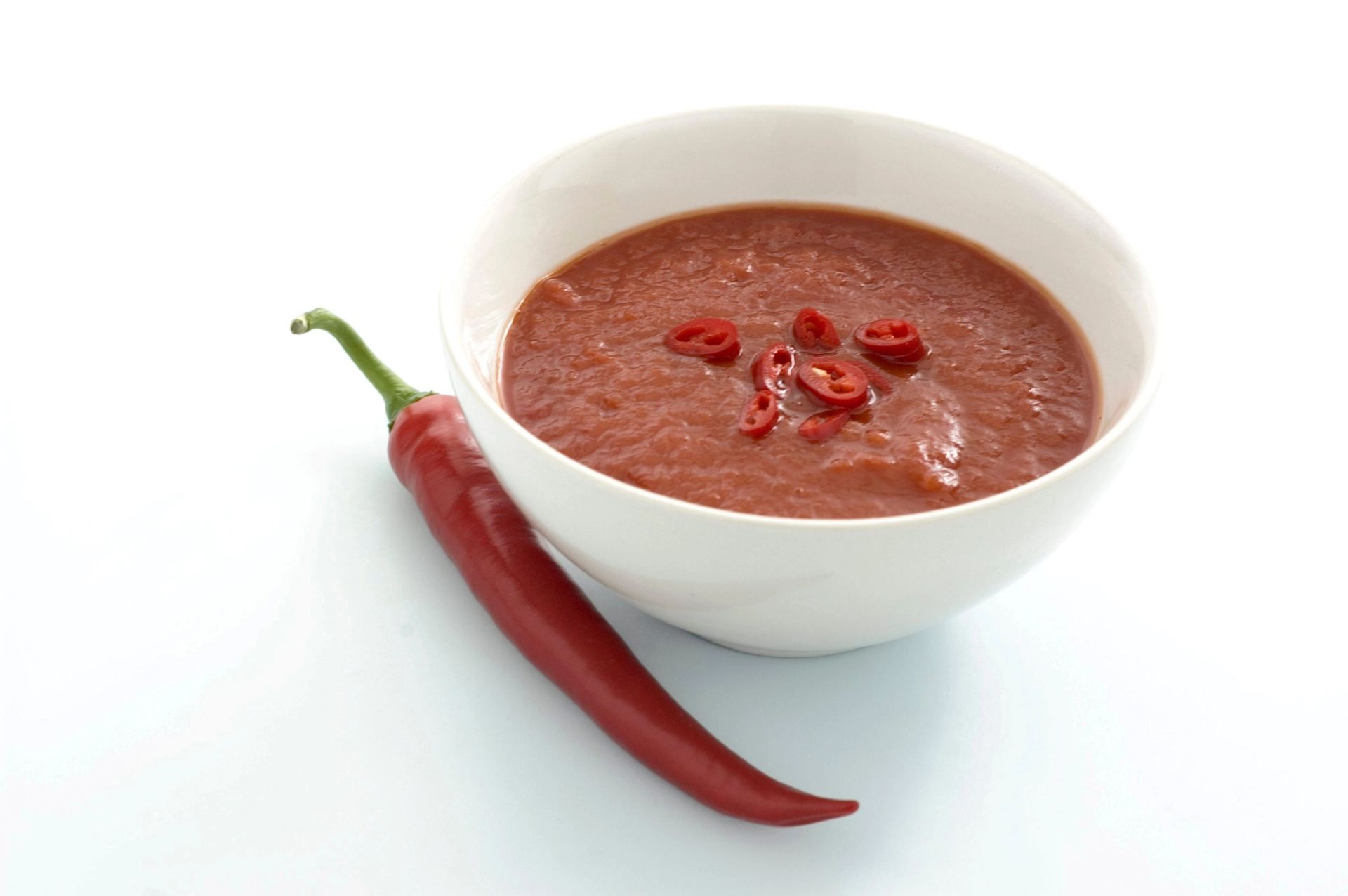 En skål med rød chilisaus toppet med skiver av chili, plassert ved siden av en hel rød chilipepper på en lys bakgrunn.