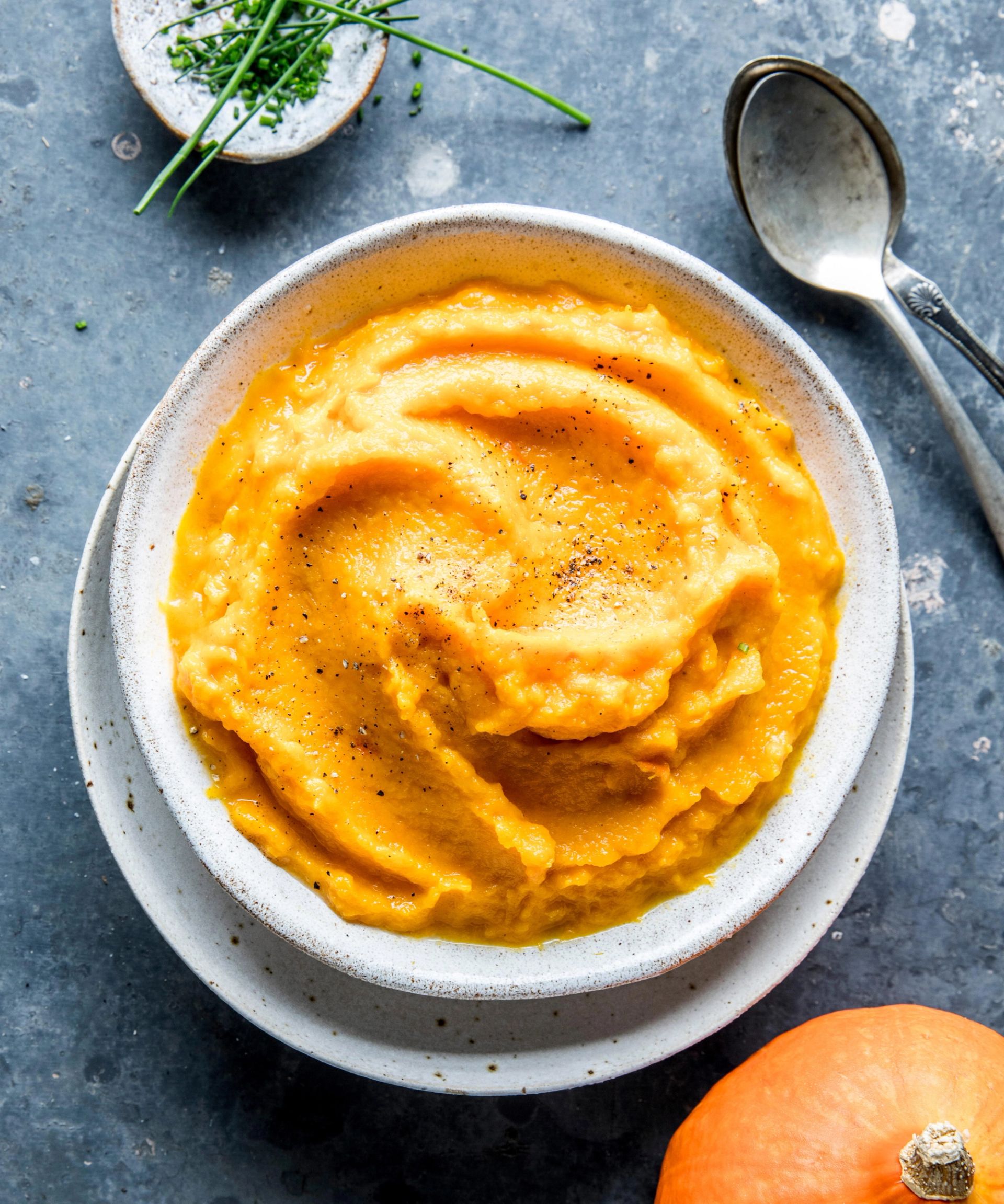En lys grå skål fylt med kremet, oransje gresskarpuré, drysset med svart pepper. Skålen står på en tallerken på en mørk steinoverflate, med en skje ved siden av og ferske urter og et helt gresskar delvis synlig i hjørnet.
