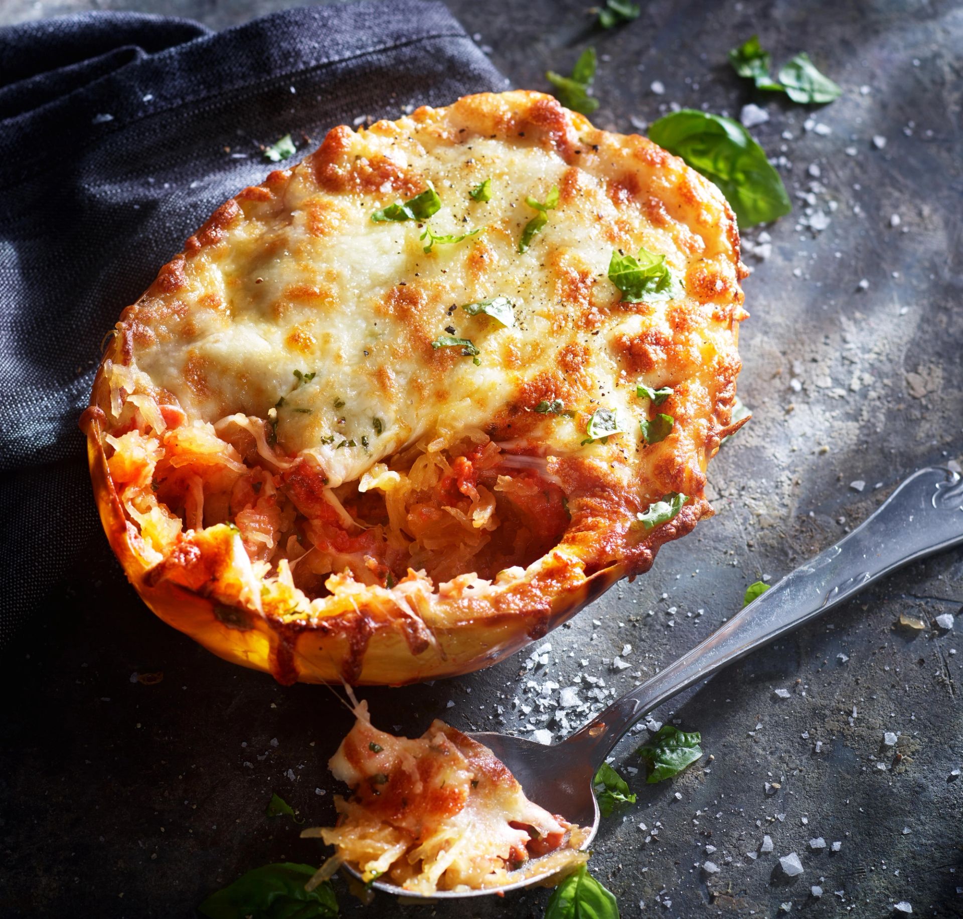 Et bakt spagettigresskar brukes som form og er fylt med en gratinert rett toppet med smeltet ost og urter. En skje ligger foran og viser litt av den myke, trådlignende gresskarmassen og fyllet. Retten står på en mørk overflate med et kjøkkenhåndkle i bakgrunnen.
