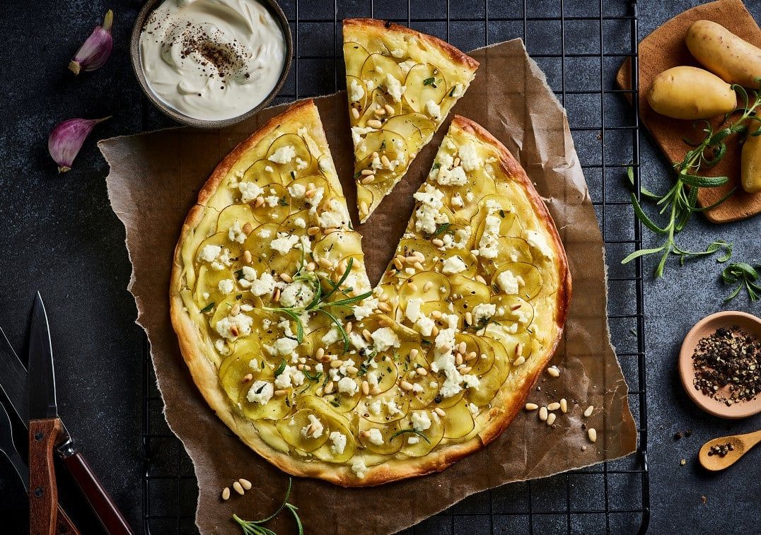 En nystekt hvit pizza toppet med tynne, overlappende potetskiver, smuldret fetaost, pinjekjerner og frisk rosmarin.