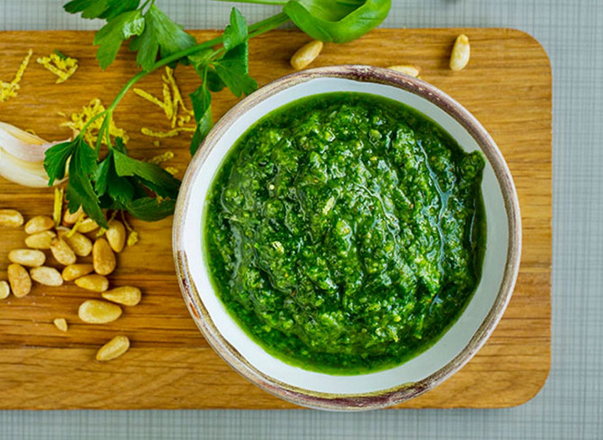 Skål med frisk, grønn pesto plassert på en treplate, omgitt av pinjekjerner, persilleblader, hvitløk og revet sitronskall.