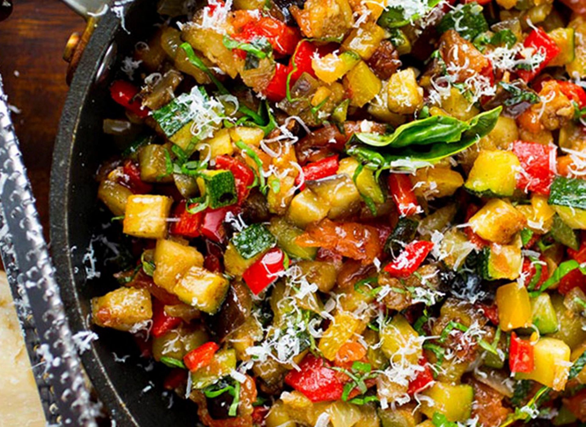 Nærbilde av en stekepanne fylt med finhakkede, stekte grønnsaker som aubergine, paprika og squash, toppet med friske urter og revet ost.