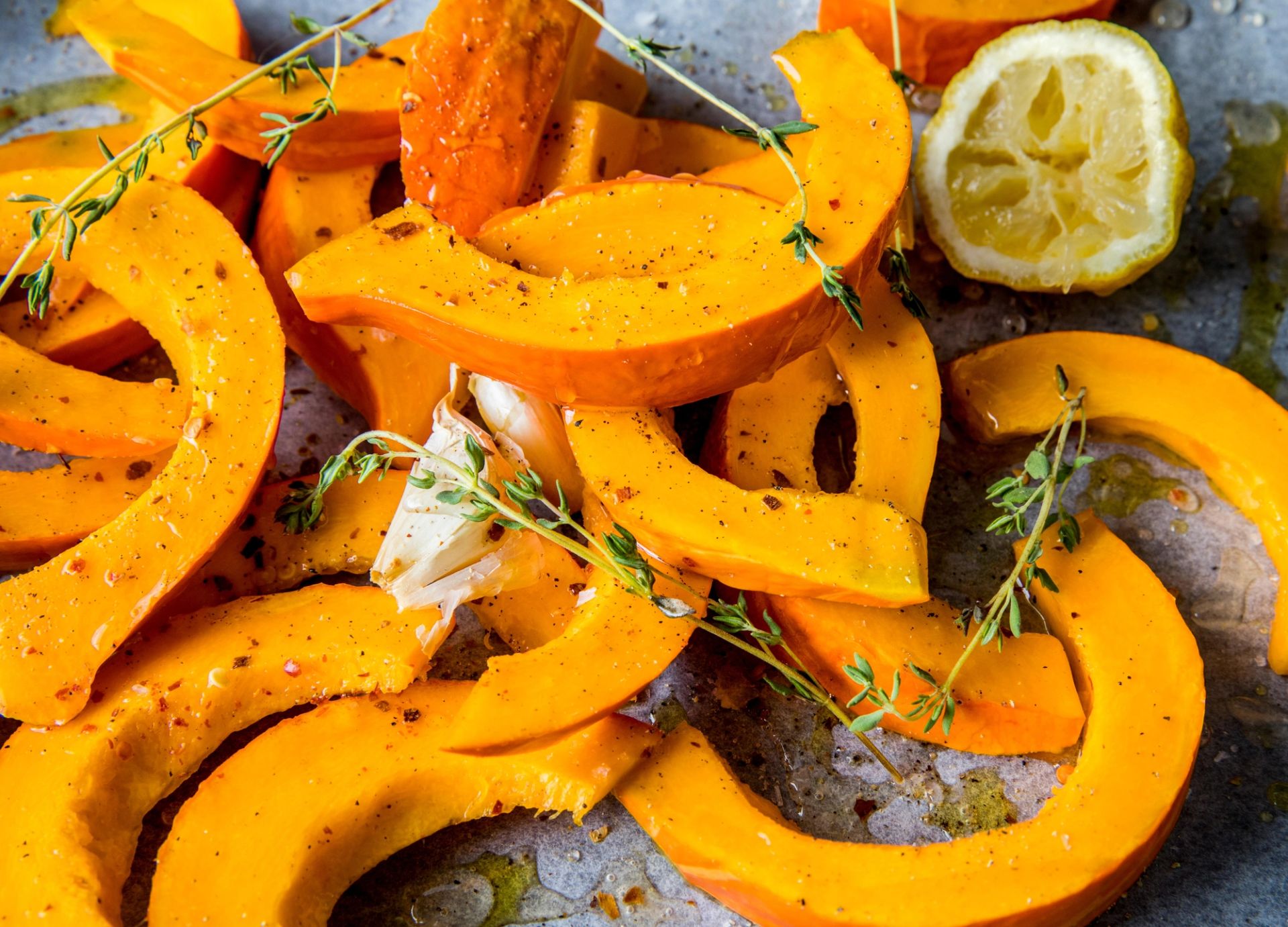 Ovnsbakte skiver av gresskar eller butternut-squash ligger spredt på et stekebrett, krydret med pepper, urter og olivenolje, med friske timiankvister, hvitløksfedd og en delt sitron synlig blant de gyllenoransje bitene.