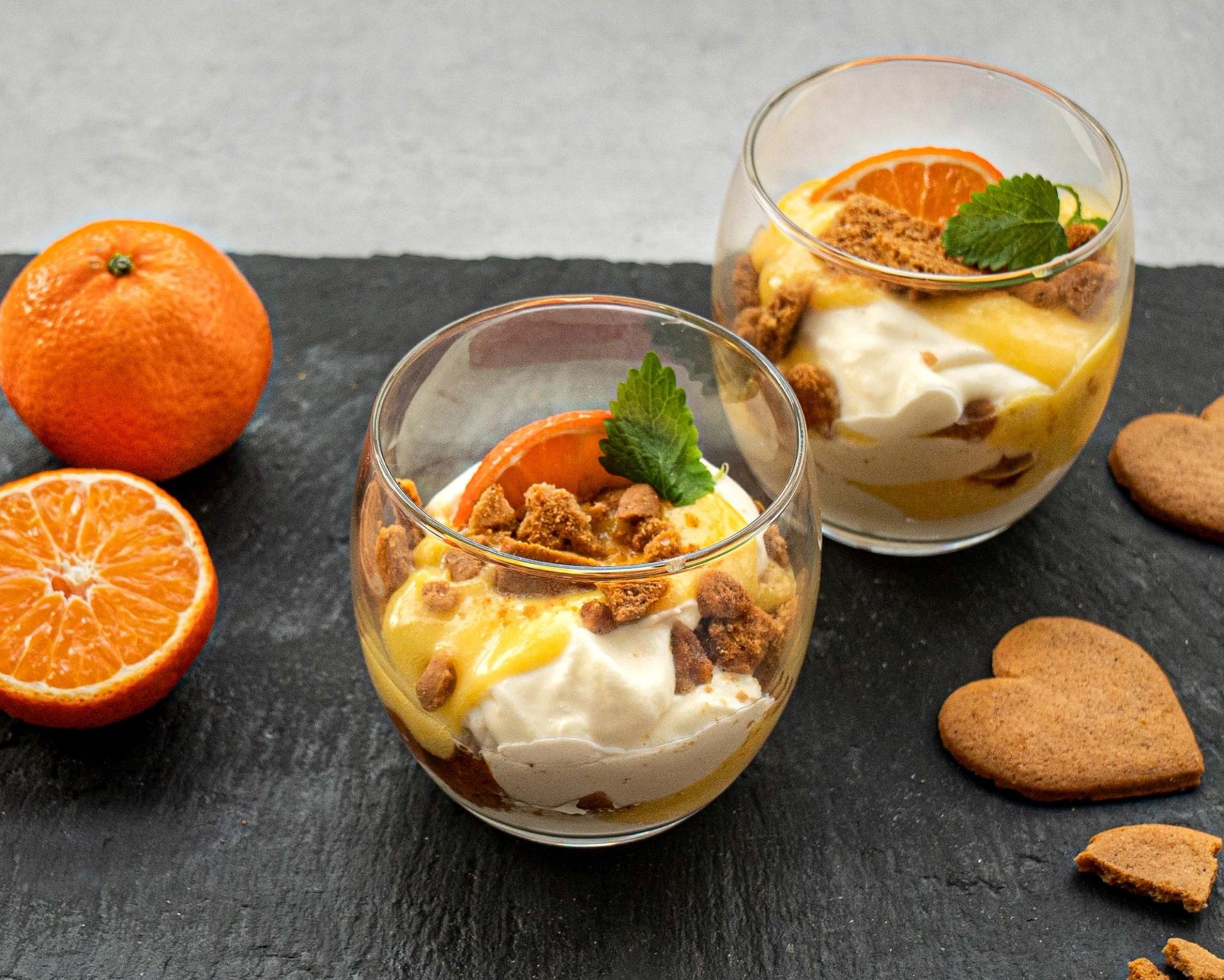 To dessertglass fylt med klementin curd, knuste pepperkaker og ostekrem, pyntet med klementinskive og sitronmelisse, står på en mørk skiferplate dekorert med med klementiner og pepperkakehjerter..