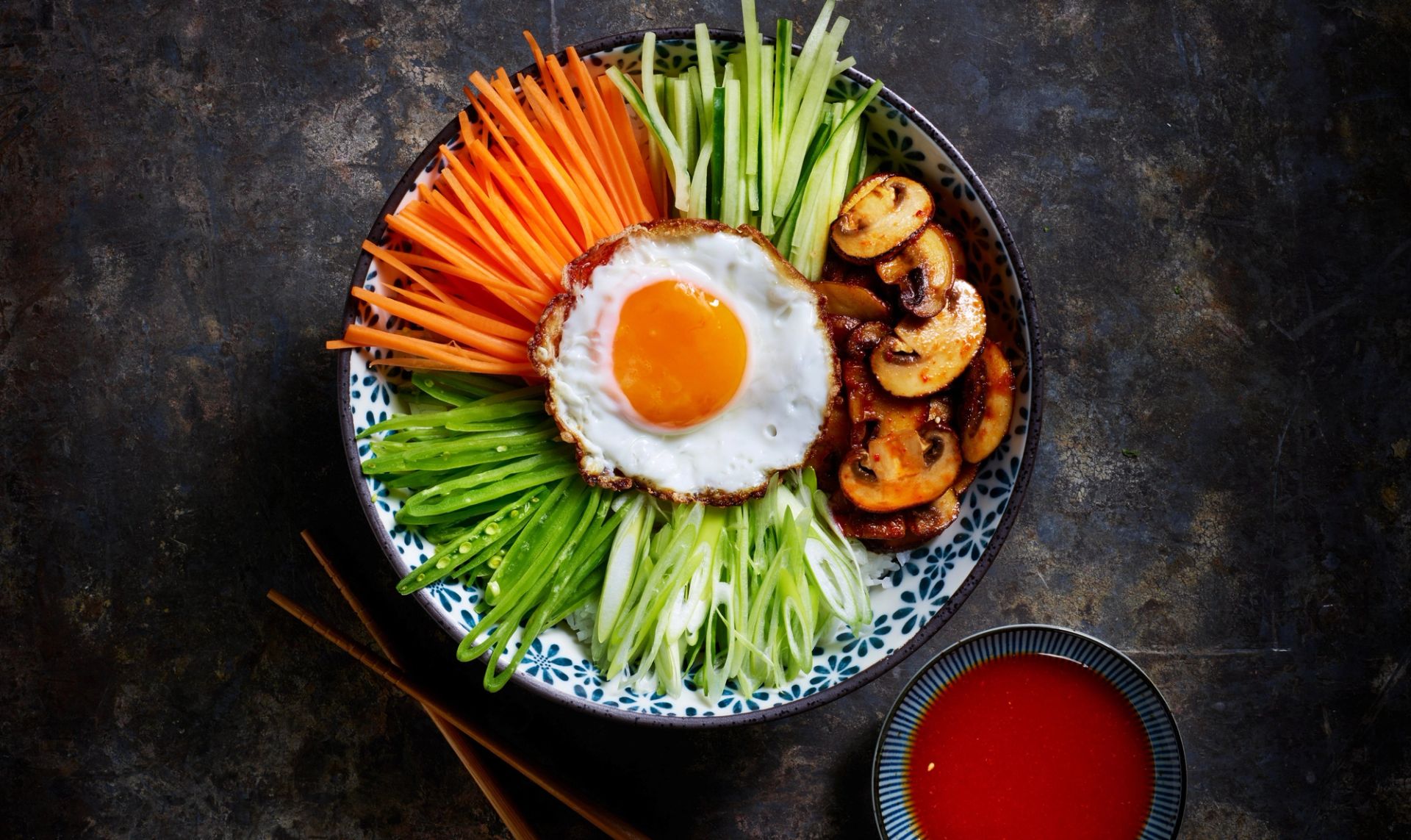 En fargerik bibimbap-bolle prydet med et stekt speilegg i midten, omgitt av seksjoner med finsnittet gulrot, agurk, grønn chili og vårløk, samt stekt sopp.