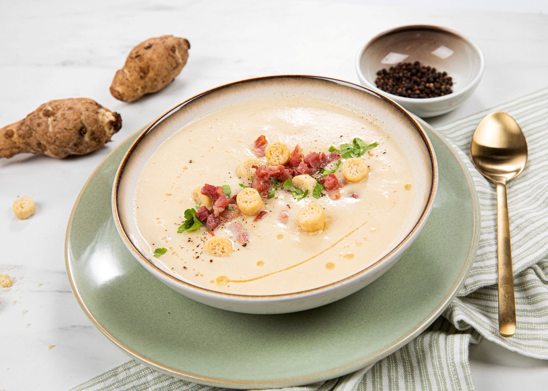 Kremet jordskokksuppe servert i en dyp tallerken, toppet med sprøstekte baconbiter, urter og sprø krutonger. Suppen er pyntet med noen dråper olje og står på en grønn tallerken, omgitt av hele jordskokker, en skål med pepper og en gyllen skje på et lyst bord.