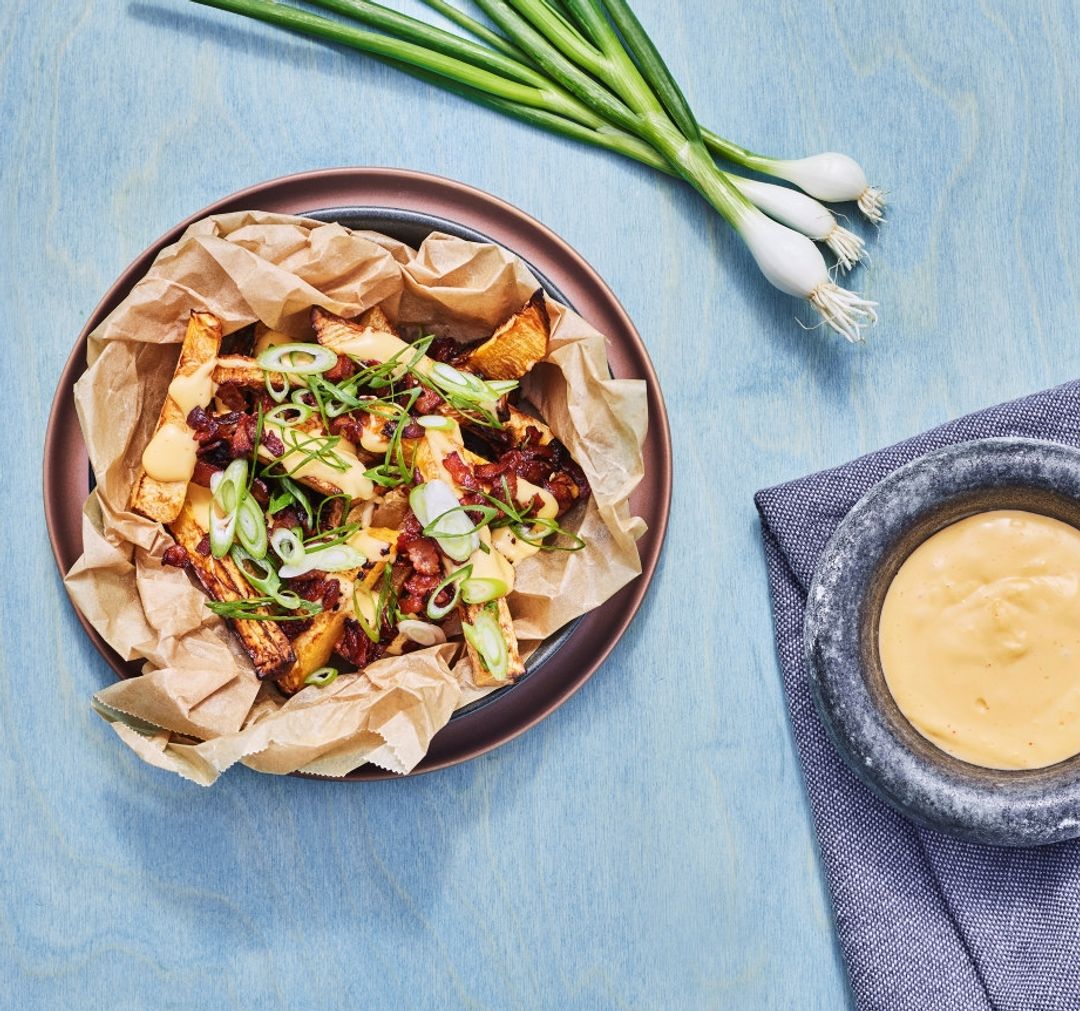 En tallerken med sprø kålrotfries toppet med sprøstekt bacon og skiver av vårløk, servert på bakepapir. Ved siden av står en liten bolle med kremet cheddarsaus. Rundt tallerkenen ligger en bunt vårløk, og underlaget er en lys blå bordflate med en brettet serviett.