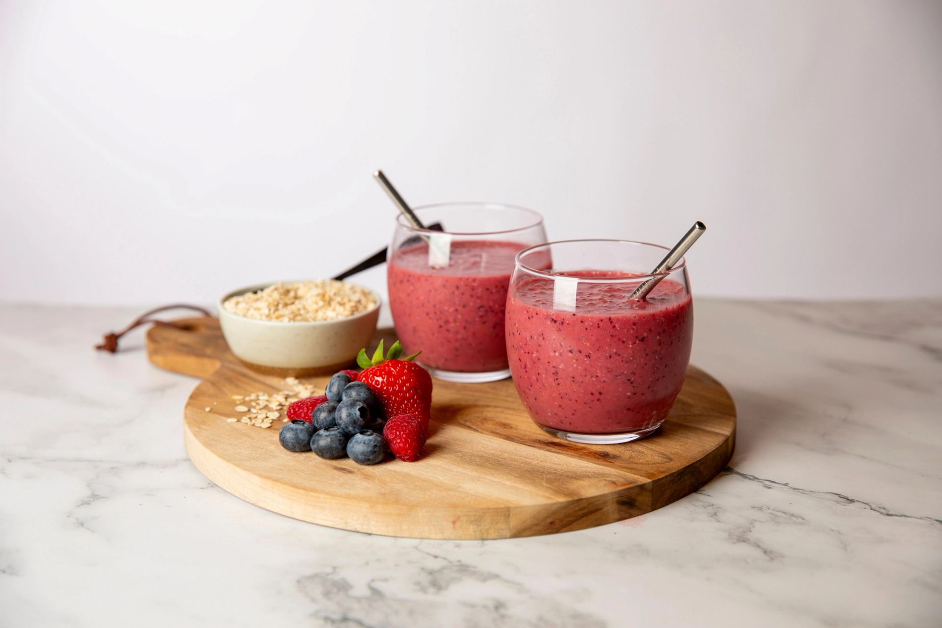 To glass med rosa smoothie av bær, servert med jordbær, blåbær og havregryn på et trefat.