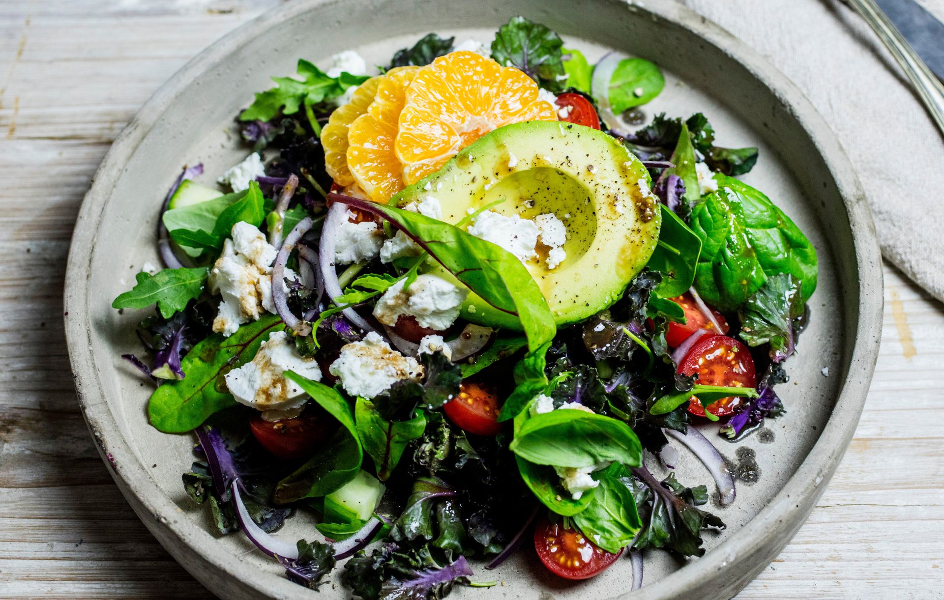 En fargerik rosettkålsalat servert i en rustikk grå bolle på et lyst trebord. Salaten er toppet med en halv avokado, skiver av appelsin, smuldret chevre, cherrytomater og rødløk, dristet med en mørk balsamicodressing.