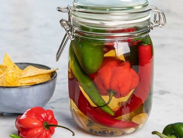 Glass med syltede chili i ulike farger, omgitt av friske chili og en liten skål tortilla­chips.