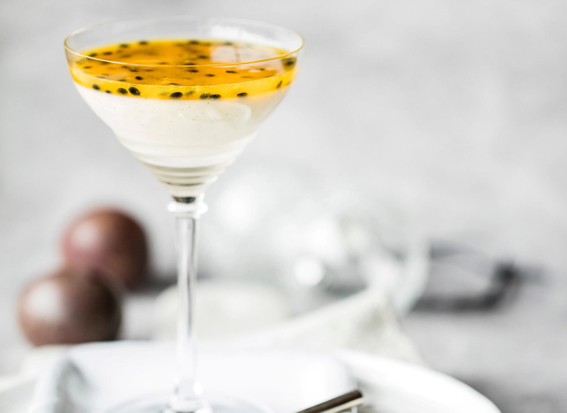 Panna cotta servert i et stettglass, toppet med gul pasjonsfruktsaus og svarte frø. Pasjonsfrukter i bakgrunnen og lys, minimalistisk bakgrunn.