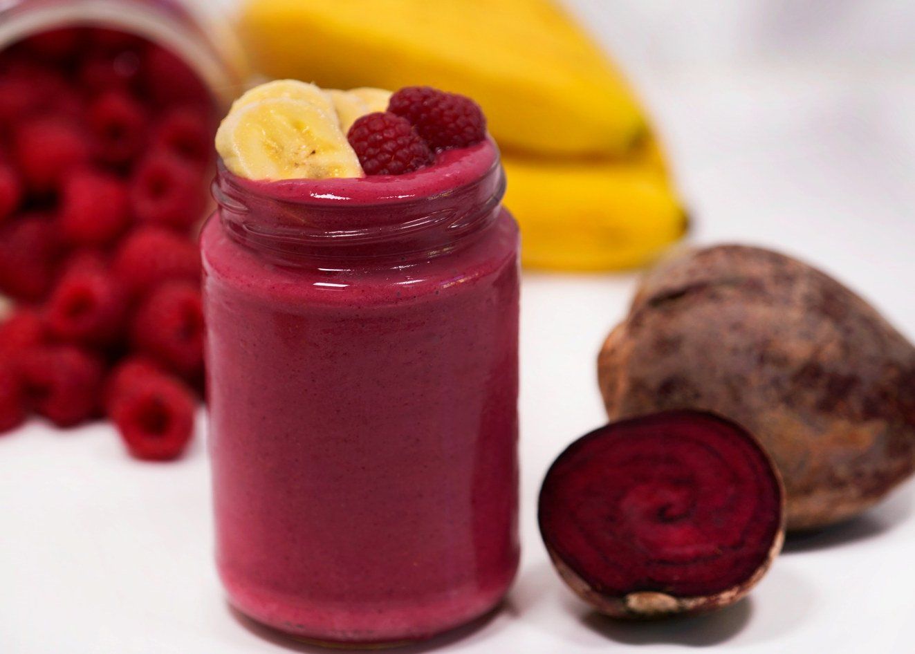 Smoothie med rødbete og bringebær servert i glass, toppet med bananskiver og friske bringebær, med bringebær og fersk rødbete i bakgrunnen.