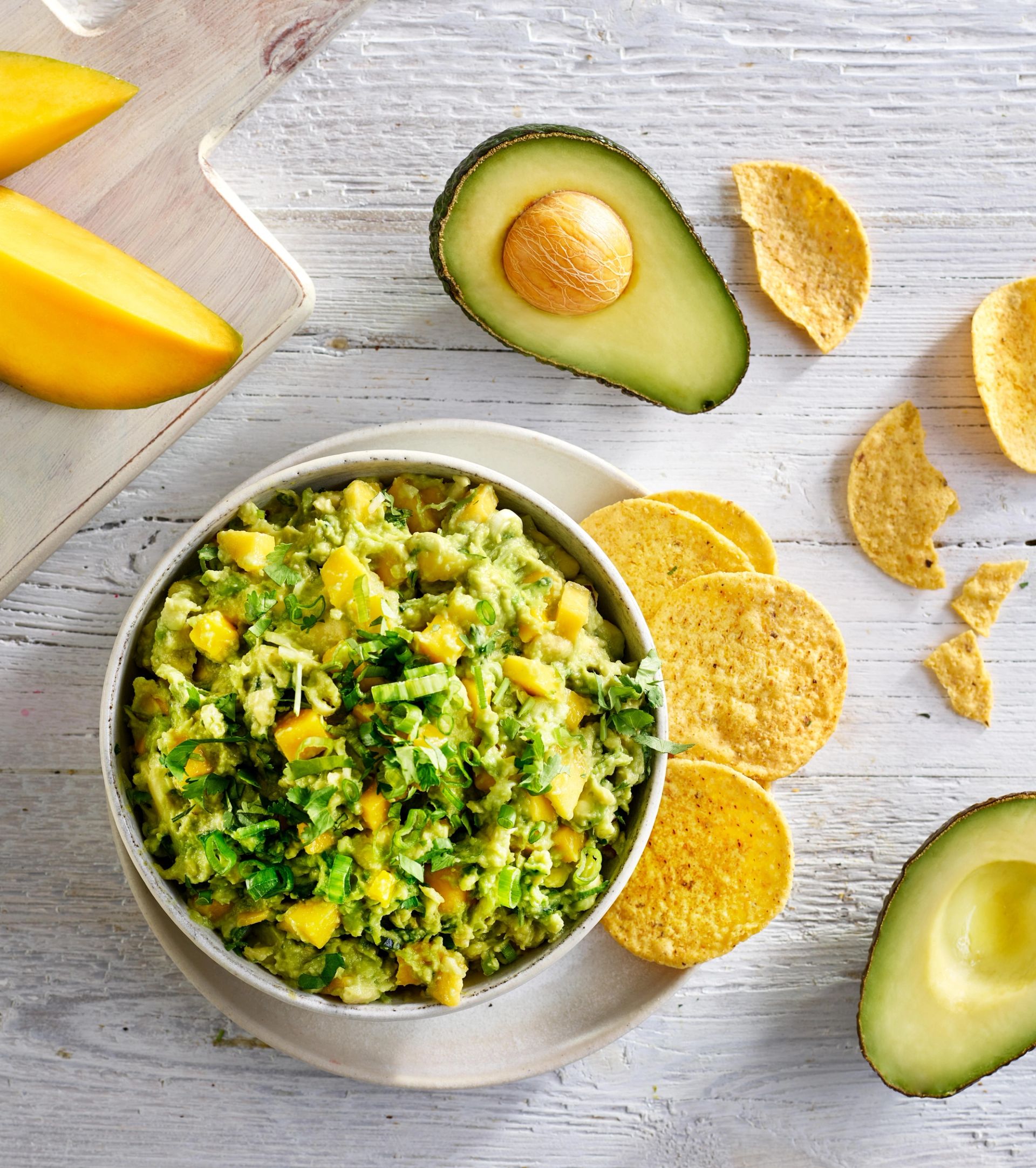 En bolle med guacamole blandet med mango og urter, omgitt av tortillachips, avokadohalvdeler og mangoskiver på et lyst, rustikt underlag.