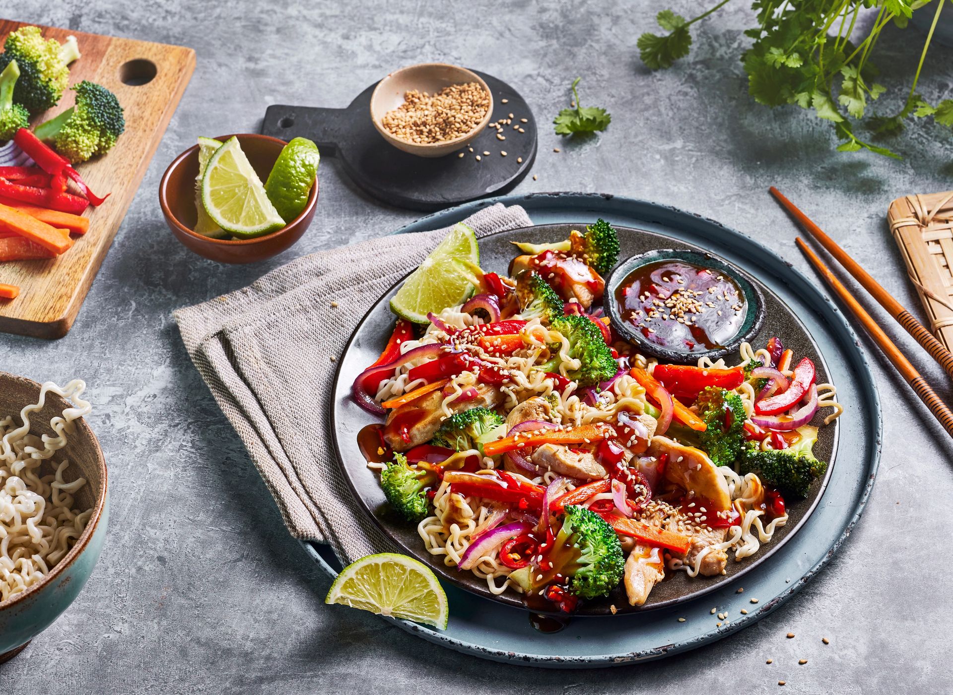 Wok med nudler, kylling, brokkoli og fargerike grønnsaker, toppet med sesamfrø, lime og saus, servert på tallerken.