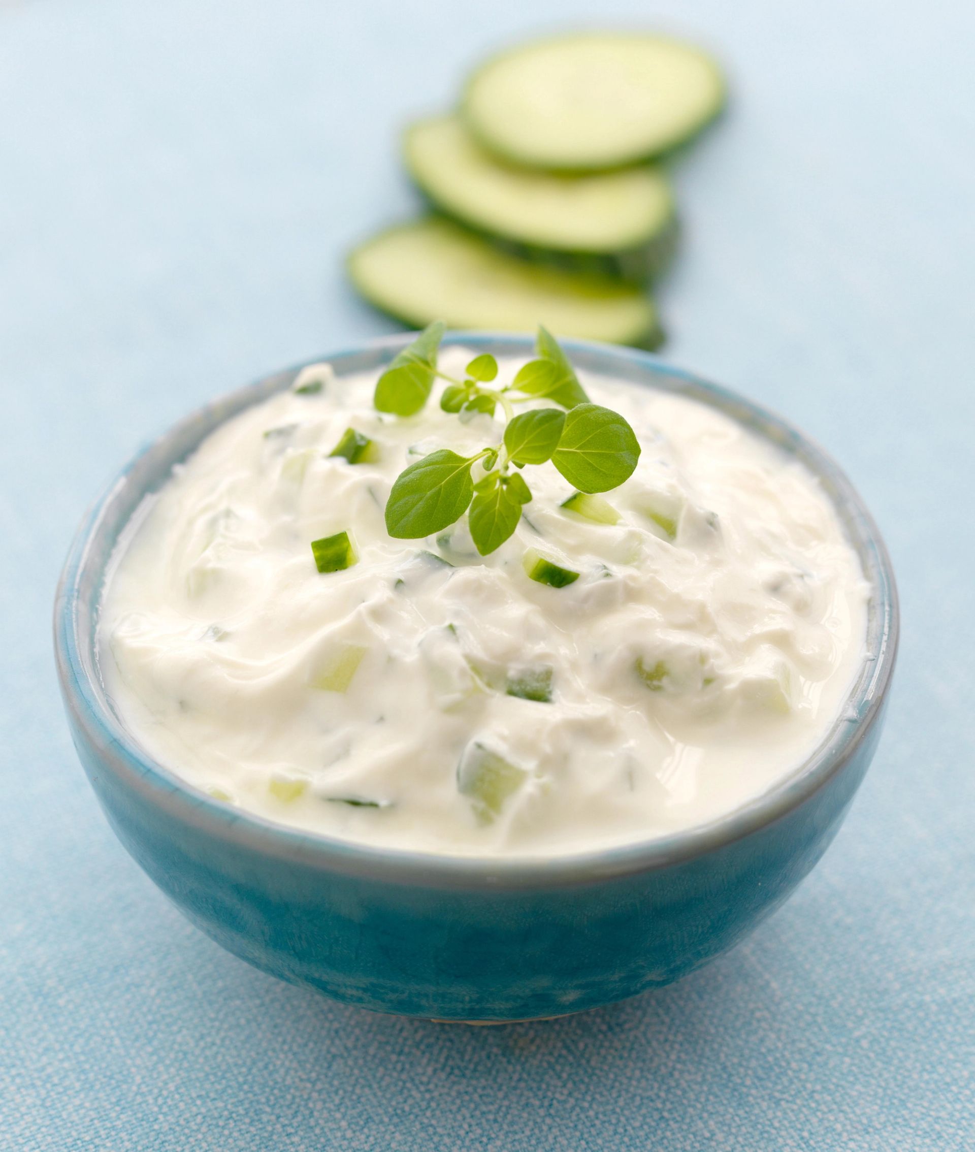 En skål med kremet tzatziki toppet med friske oregano‑blader, med skiver av agurk liggende i bakgrunnen på et lyst blått underlag.