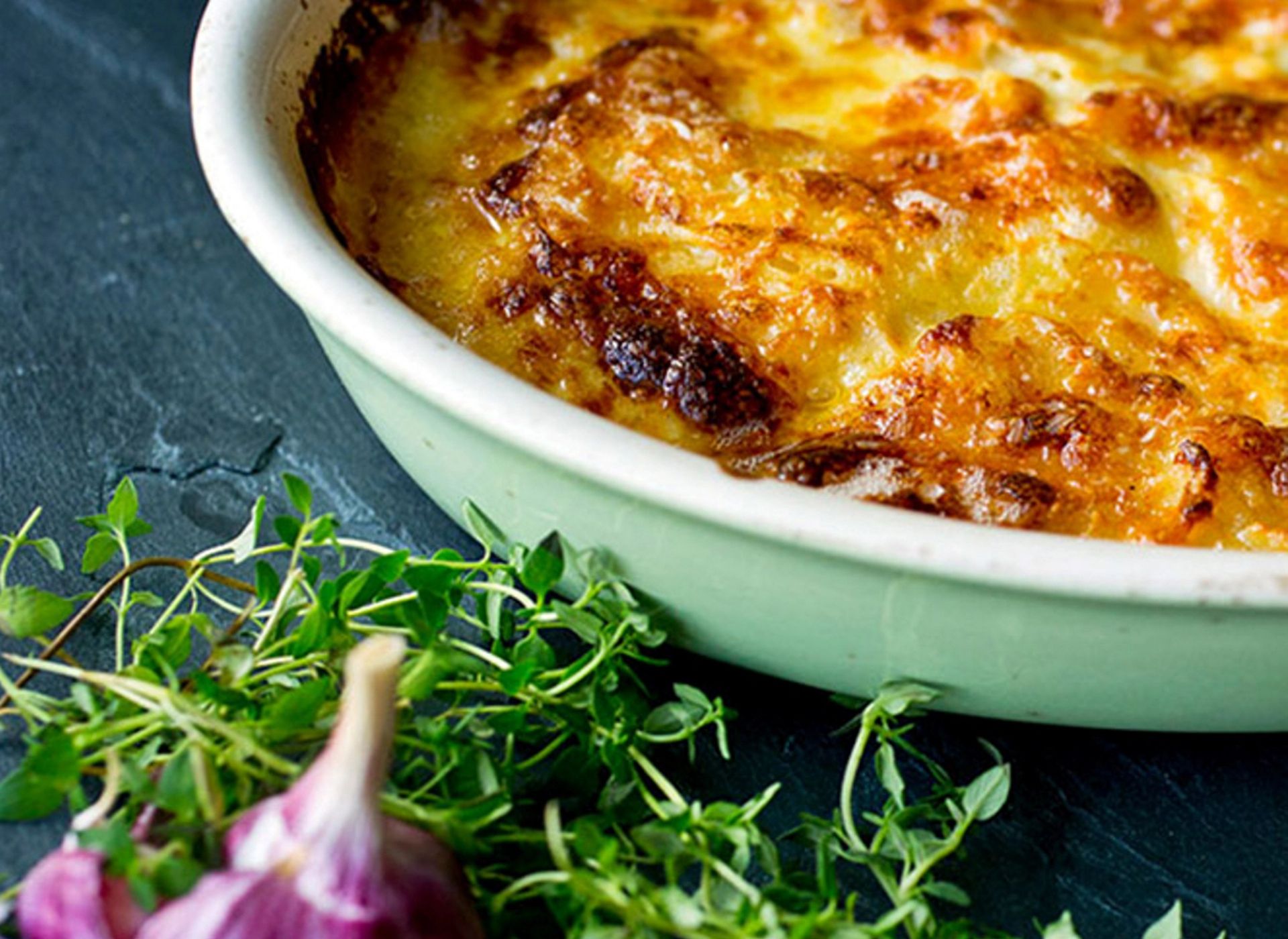 En ildfast form fylt med en ovnsbakt rett dekket av gyllen, gratinert ost med sprø, brunede topper. I forgrunnen ligger friske urter og et hvitløkshode på et mørkt underlag, som gir en rustikk kjøkkenstemning.