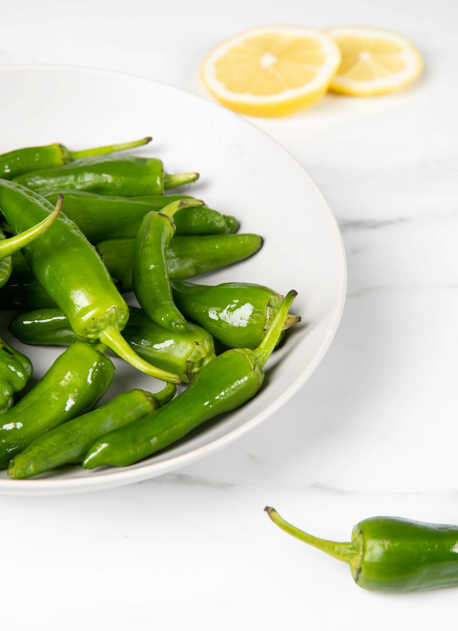 En skål med små grønne chilipepper står på et lyst bord, med sitronskiver i bakgrunnen.