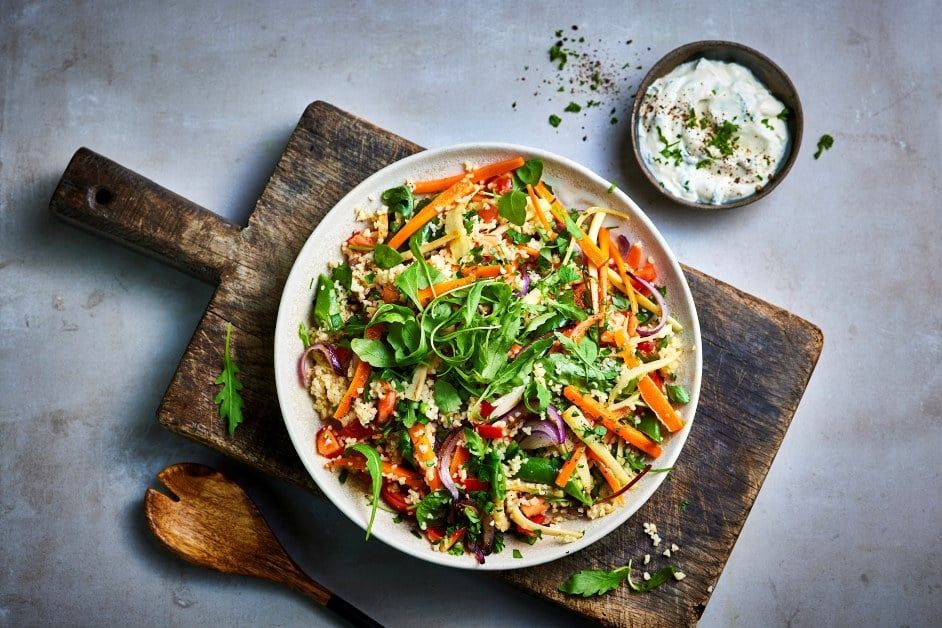 Salat med bakte rotgrønnsaker, friske grønne blader, rødløk og urter, servert i en lys bolle på et trebrett. Ved siden av står en skål med kremet dressing på grå bakgrunn.