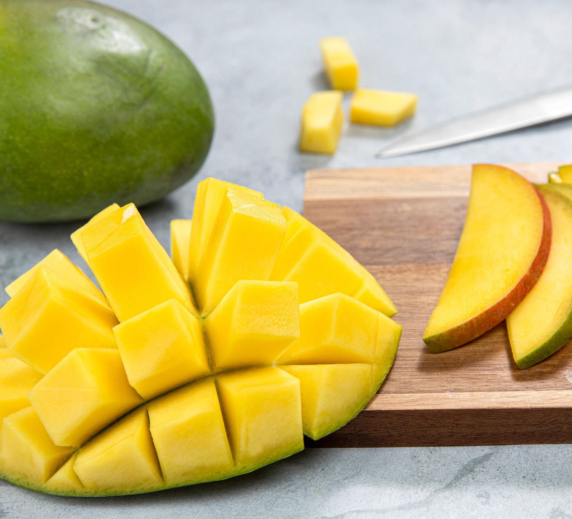 Mango skåret på to ulike måter ligger på et skjærebrett. Til venstre er en mango skåret i rutemønster og brettet ut, mens tynne mangoskiver ligger på rekke til høyre. En hel mango og en kniv er plassert i bakgrunnen på et lyst underlag.
