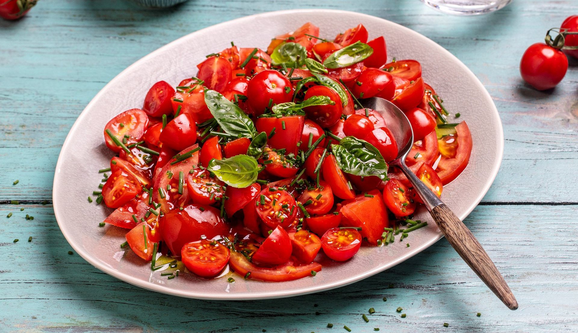 Tomatsalat med røde tomater i ulike størrelser, toppet med friske basilikumblader og finhakket gressløk, servert på et lyst fat med skje på et rustikt blåmalt trebord.