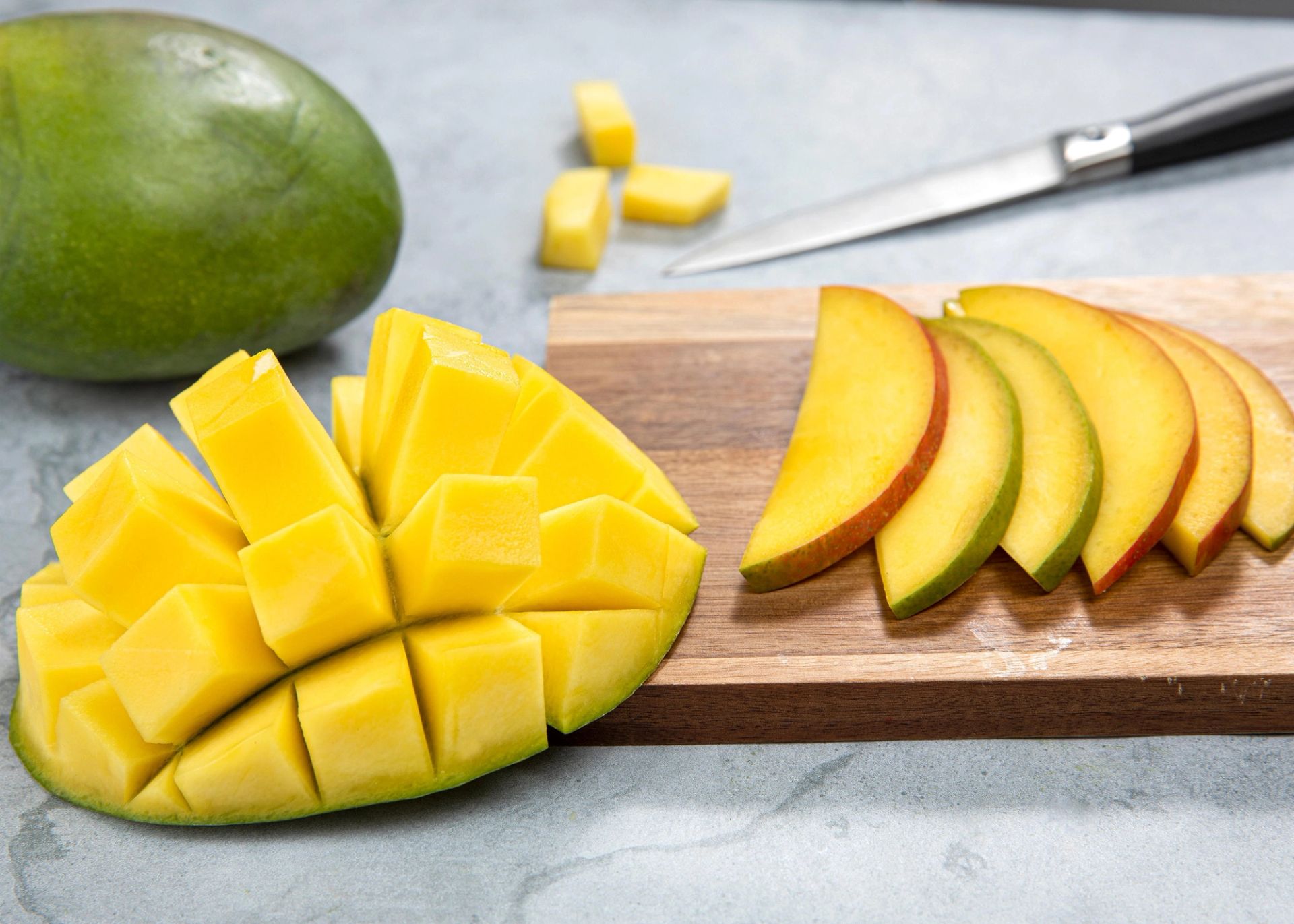 En oppskåret mango med rutemønster i fruktkjøttet ligger foran en skjærefjøl med tynne mangoskiver, sammen med en hel mango og en kniv